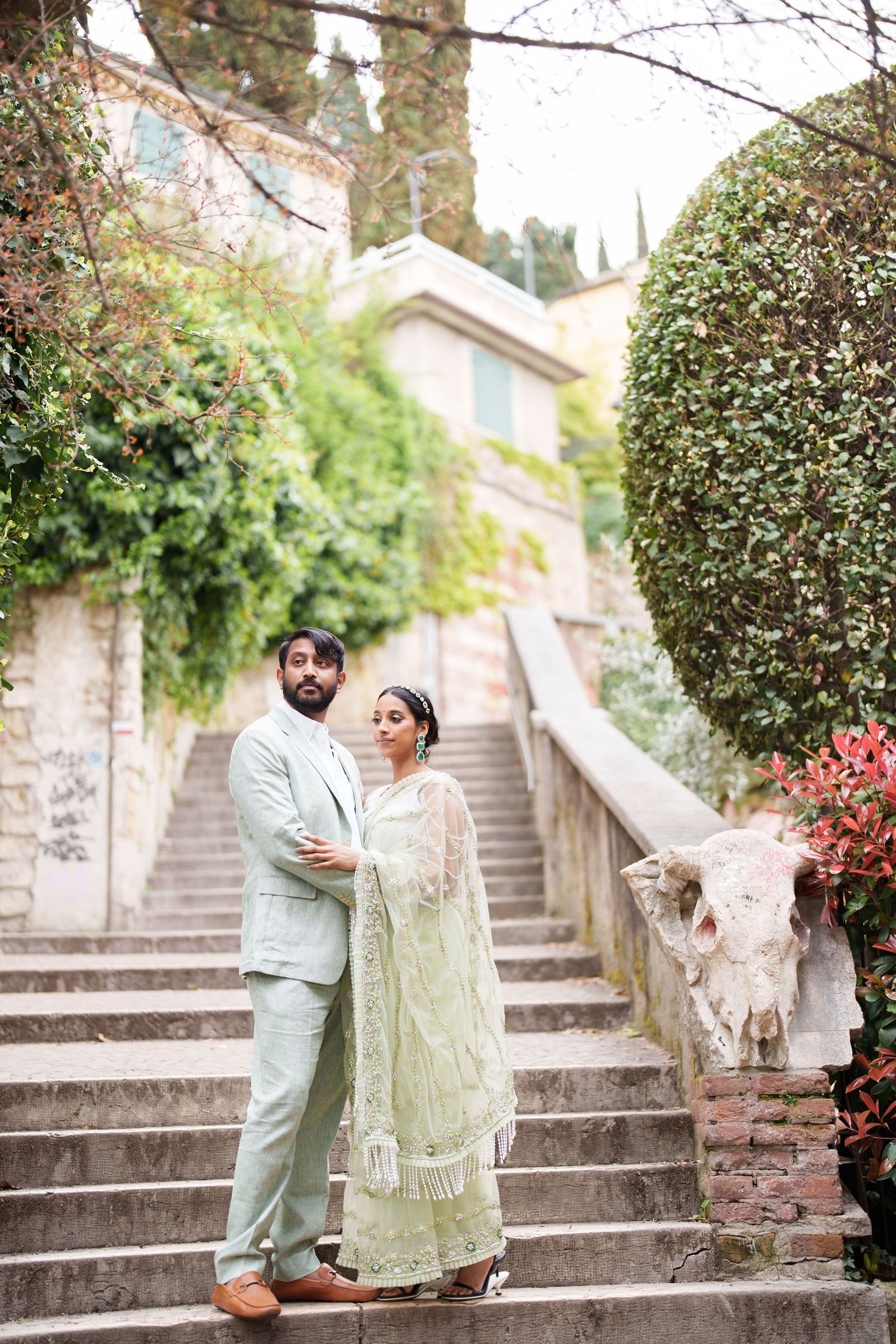 A couple in traditional South Asian attire poses on stone steps surrounded by lush greenery and flowers.