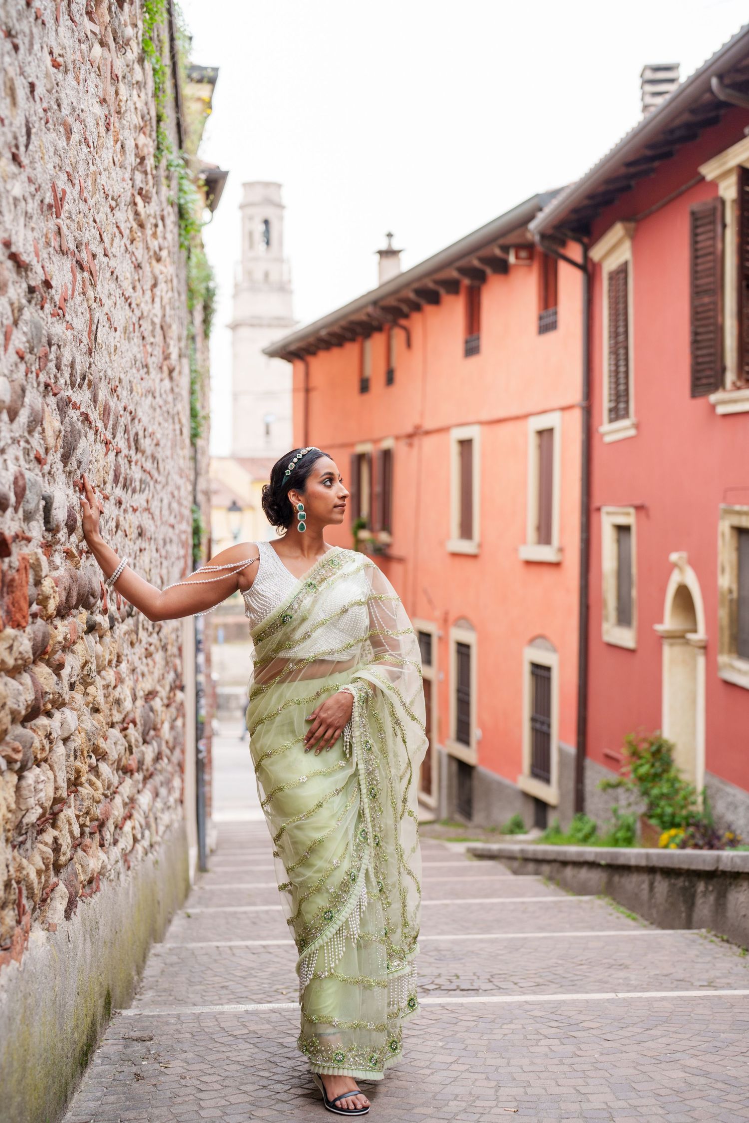 A person wearing a light green sari poses on a cobblestone street with pink buildings and a church tower in the background.