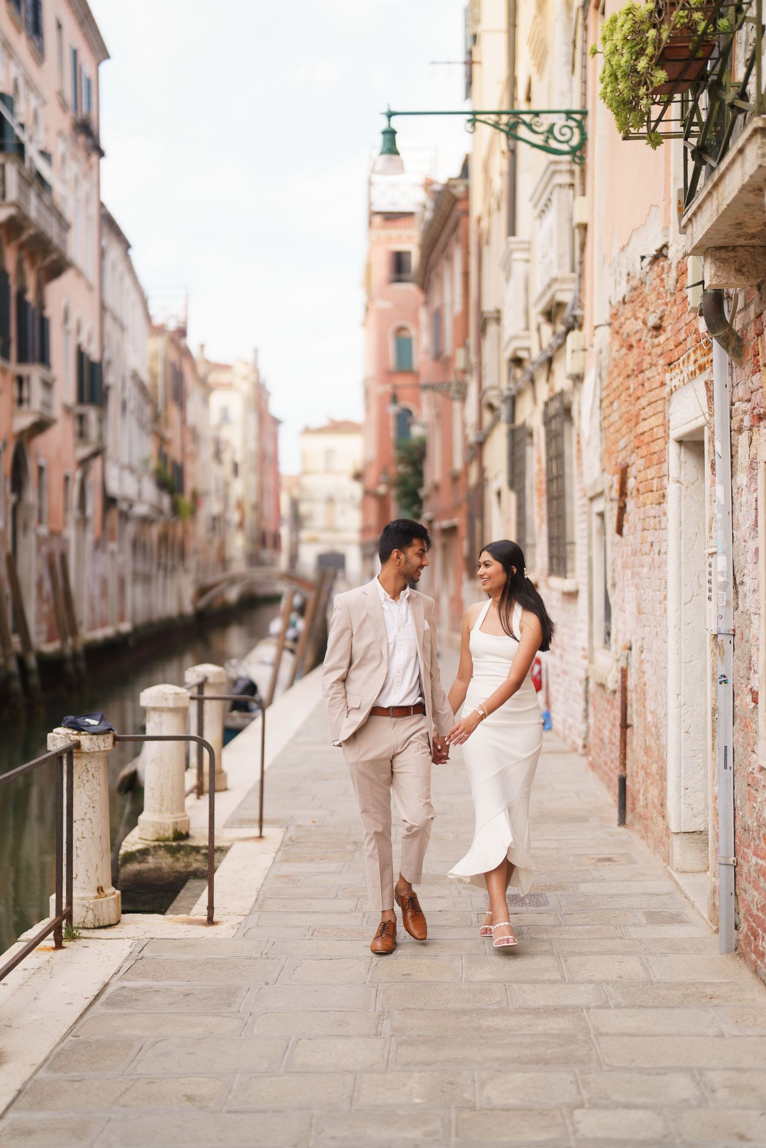 A couple strolls hand in hand along a romantic Venice canal walkway in light colored outfits.