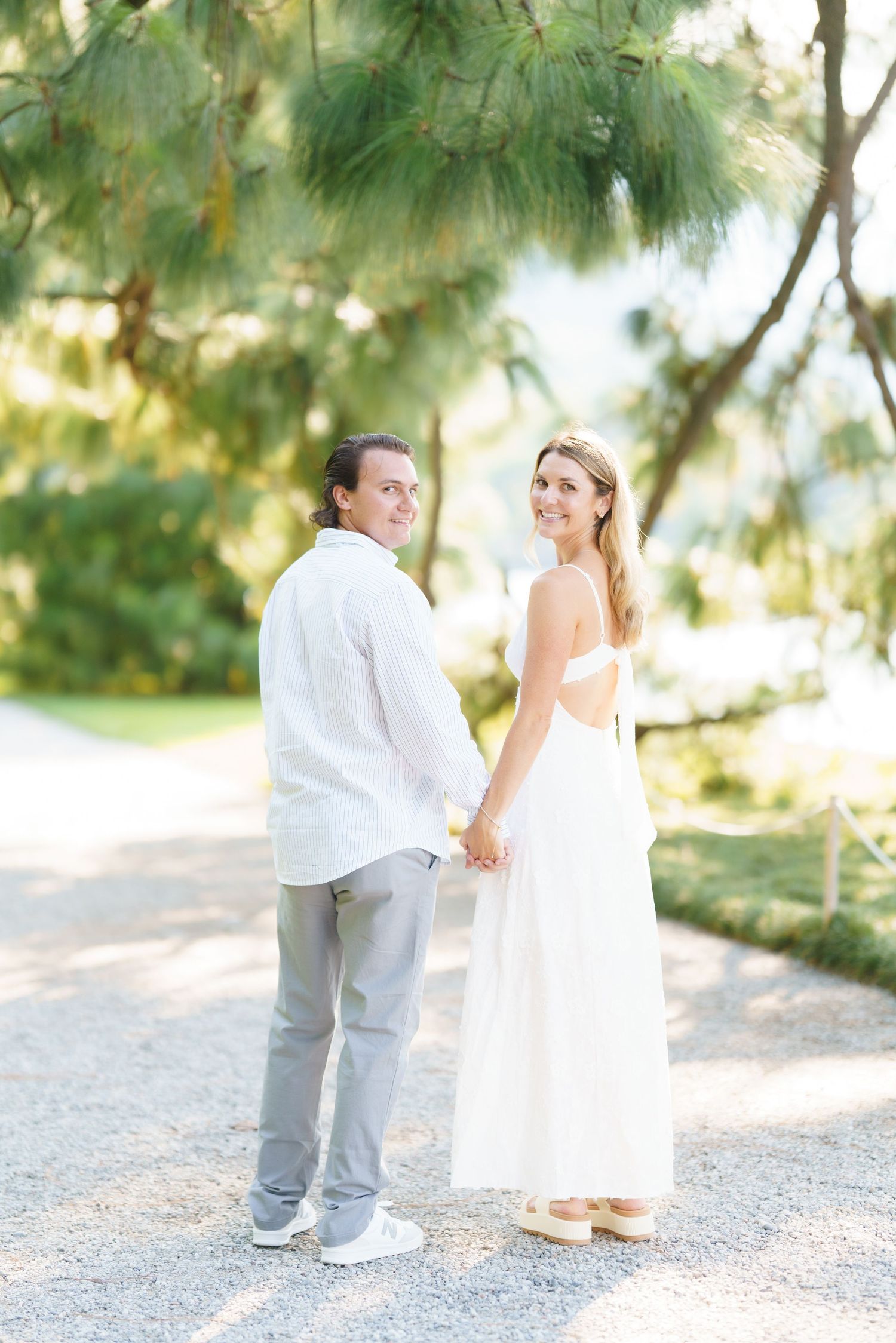 A couple in wedding attire shares a tender moment on a sunlit path lined with pine trees.