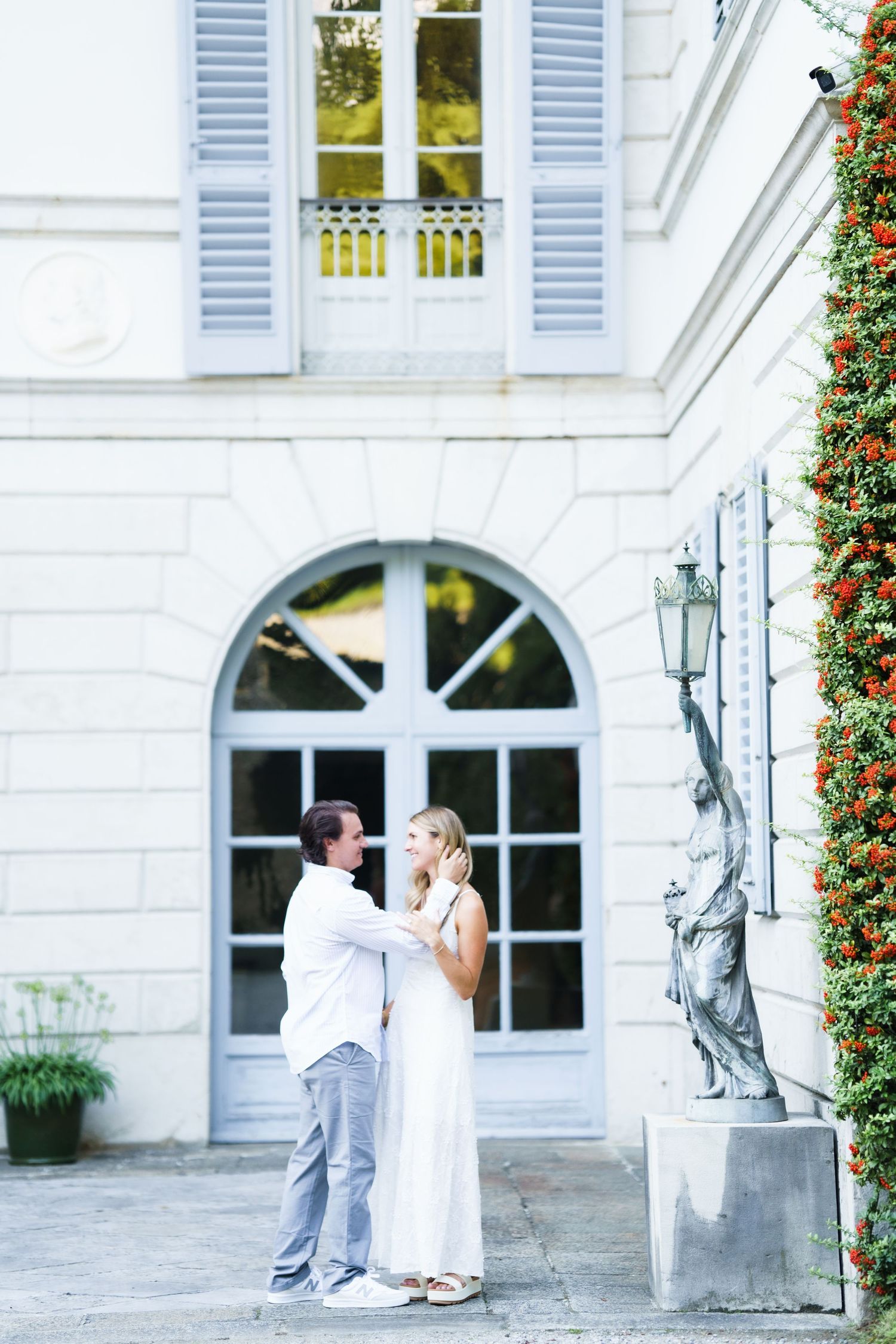 A couple shares a romantic moment outside a white mansion with blue shutters and an arched window.