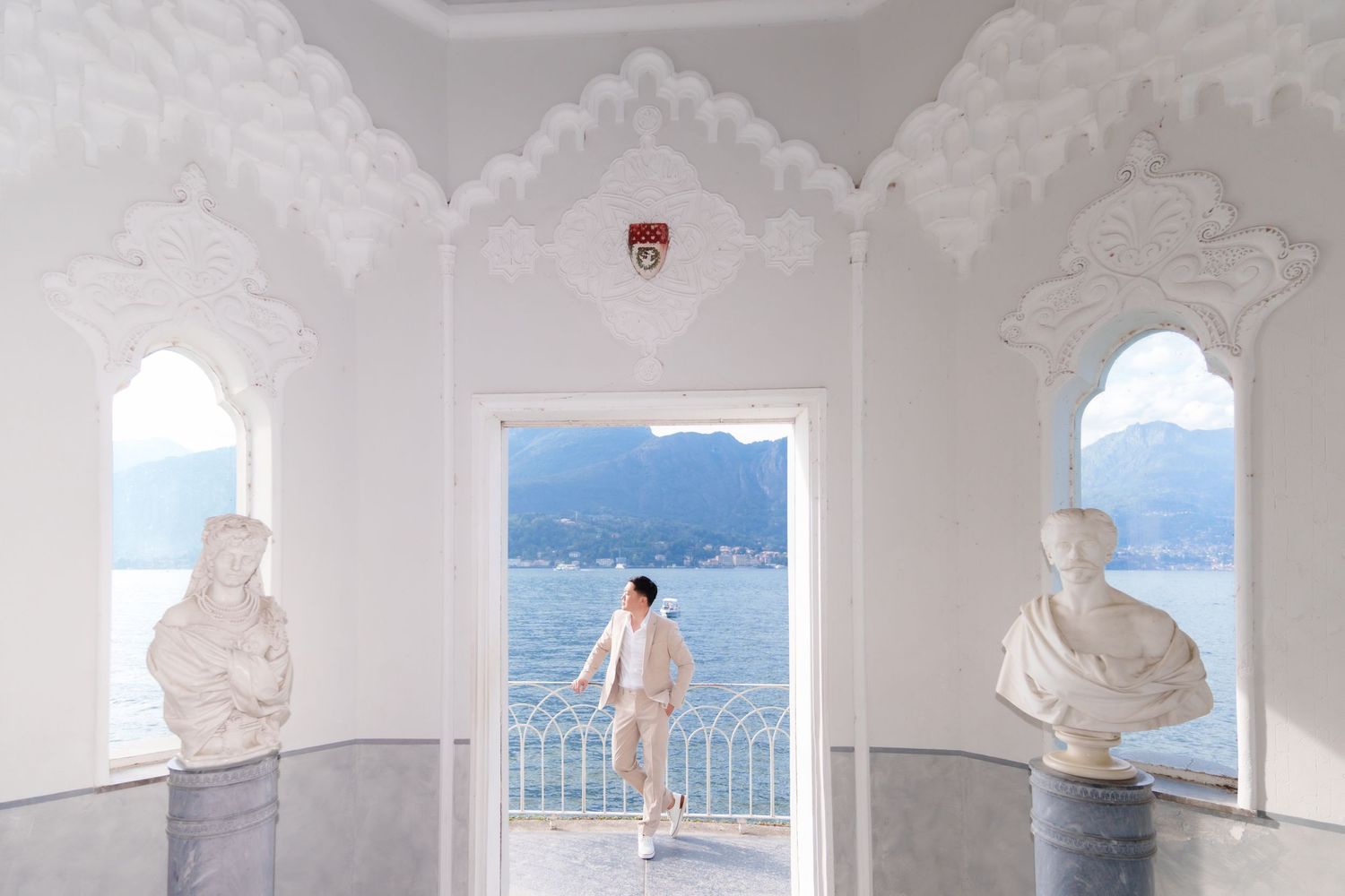 A series of arched white columns frame a scenic Mediterranean coastal view with blue water and mountains in the distance.