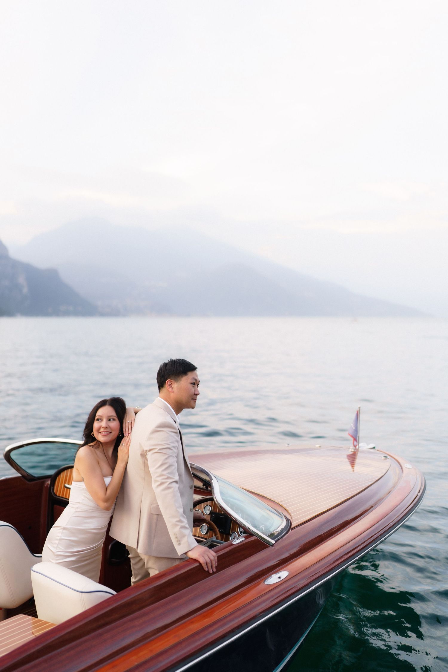 A couple enjoys a romantic boat ride on a wooden vessel against misty mountain lake backdrop.