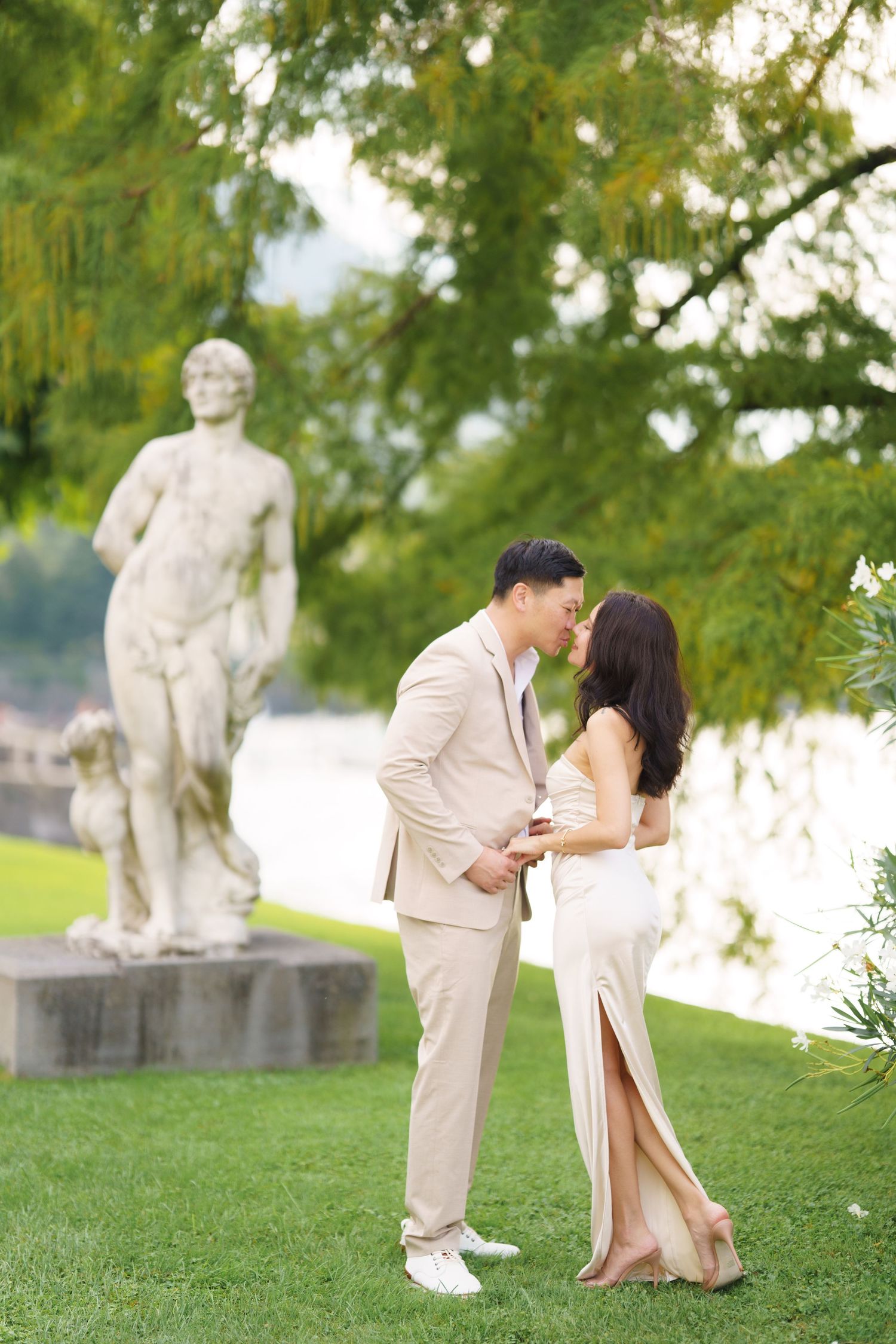 Romantic couple shares tender moments in a garden setting with classical statues and lush greenery in the background.