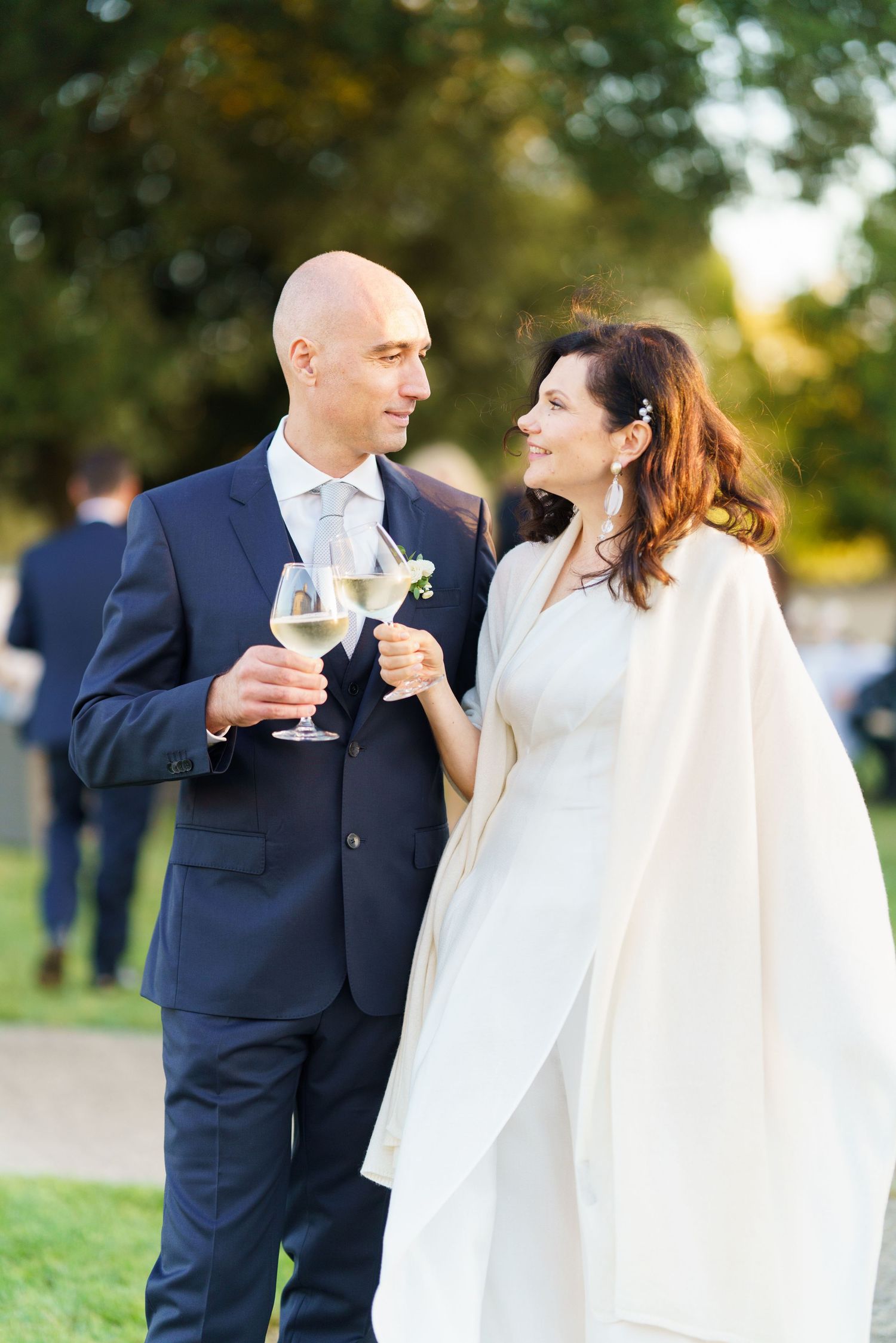 A couple in wedding attire sharing a romantic moment while holding wine glasses in an outdoor garden setting.