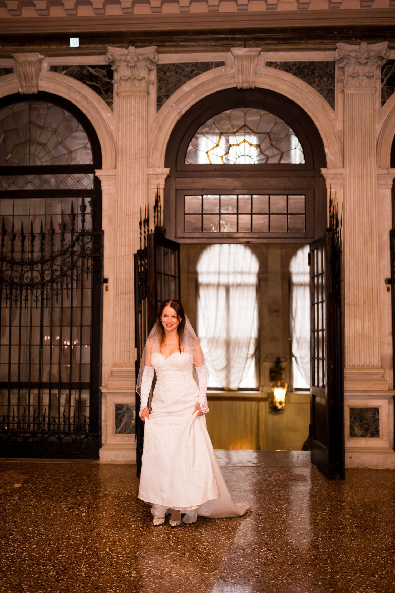 A bride in a white dress stands in an ornate stone archway with decorative ironwork gates and polished marble floors.