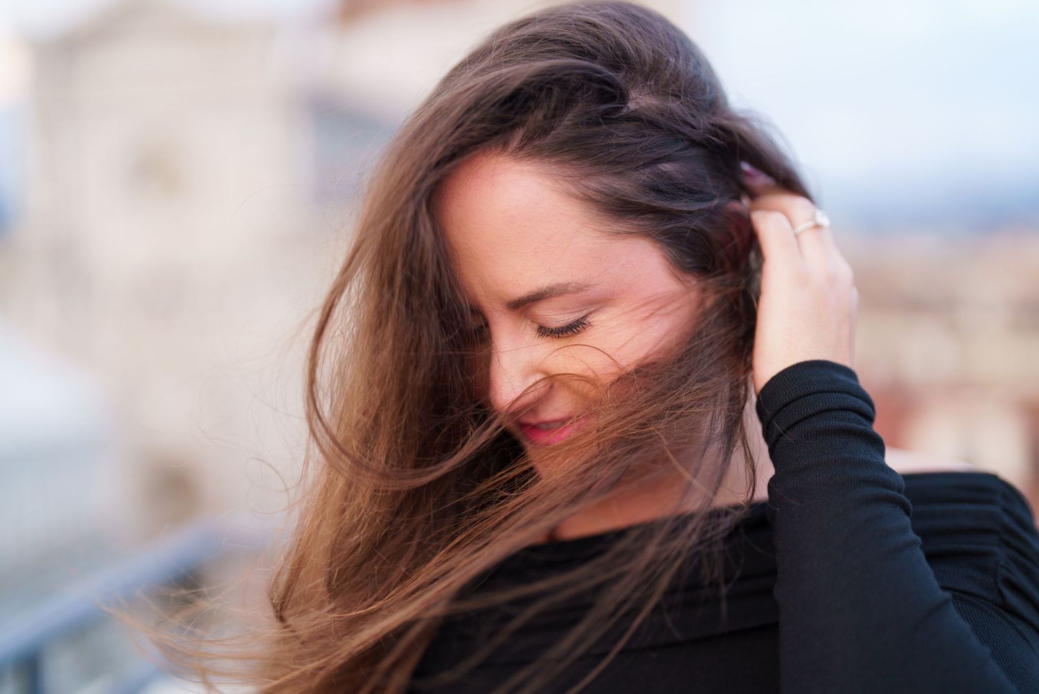 A person with long brown hair stands on a beach wearing a black sweater with mountains in the background.