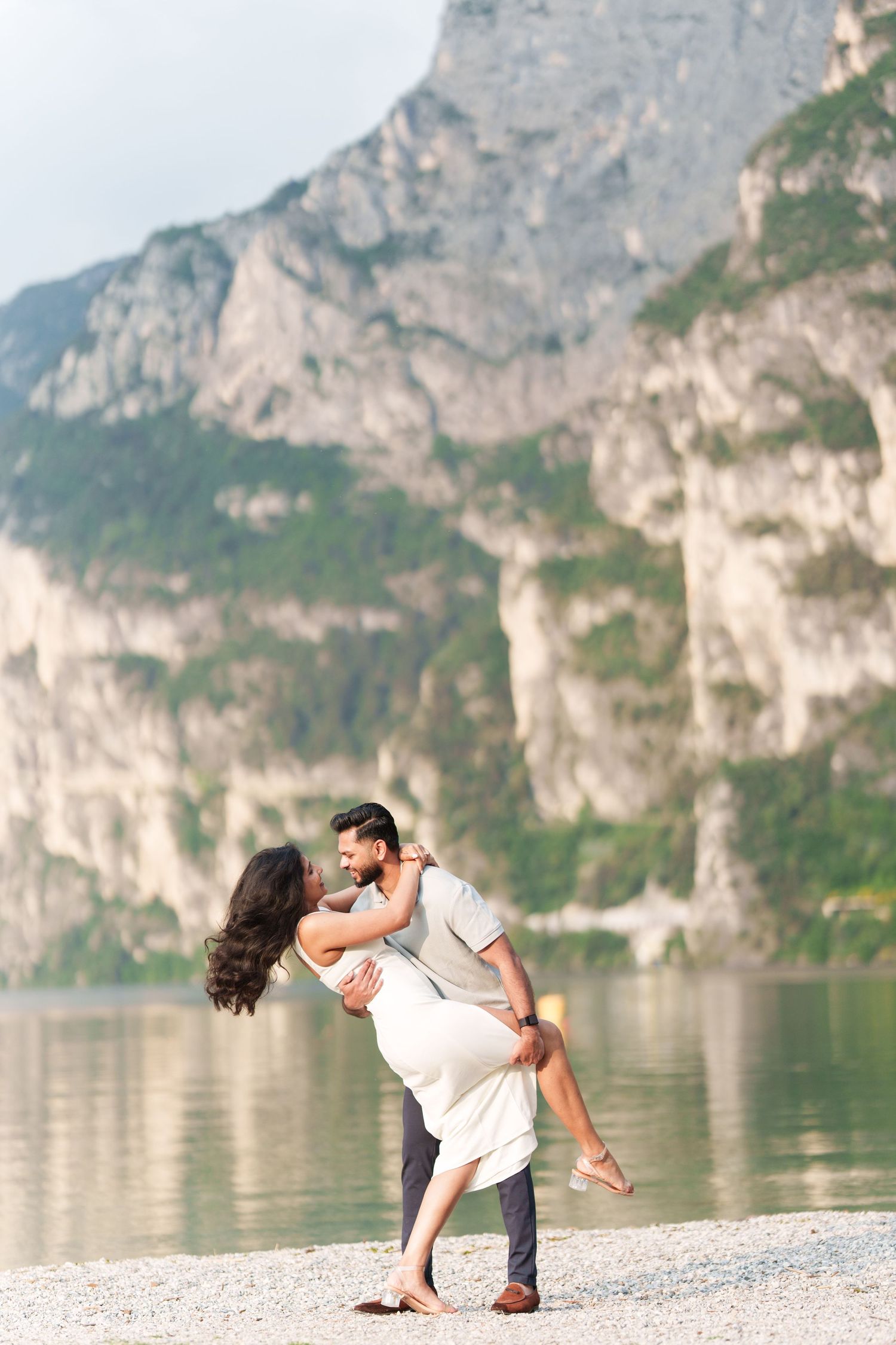 A couple performs a romantic dance pose on a beach with dramatic mountain cliffs and lake in the background.