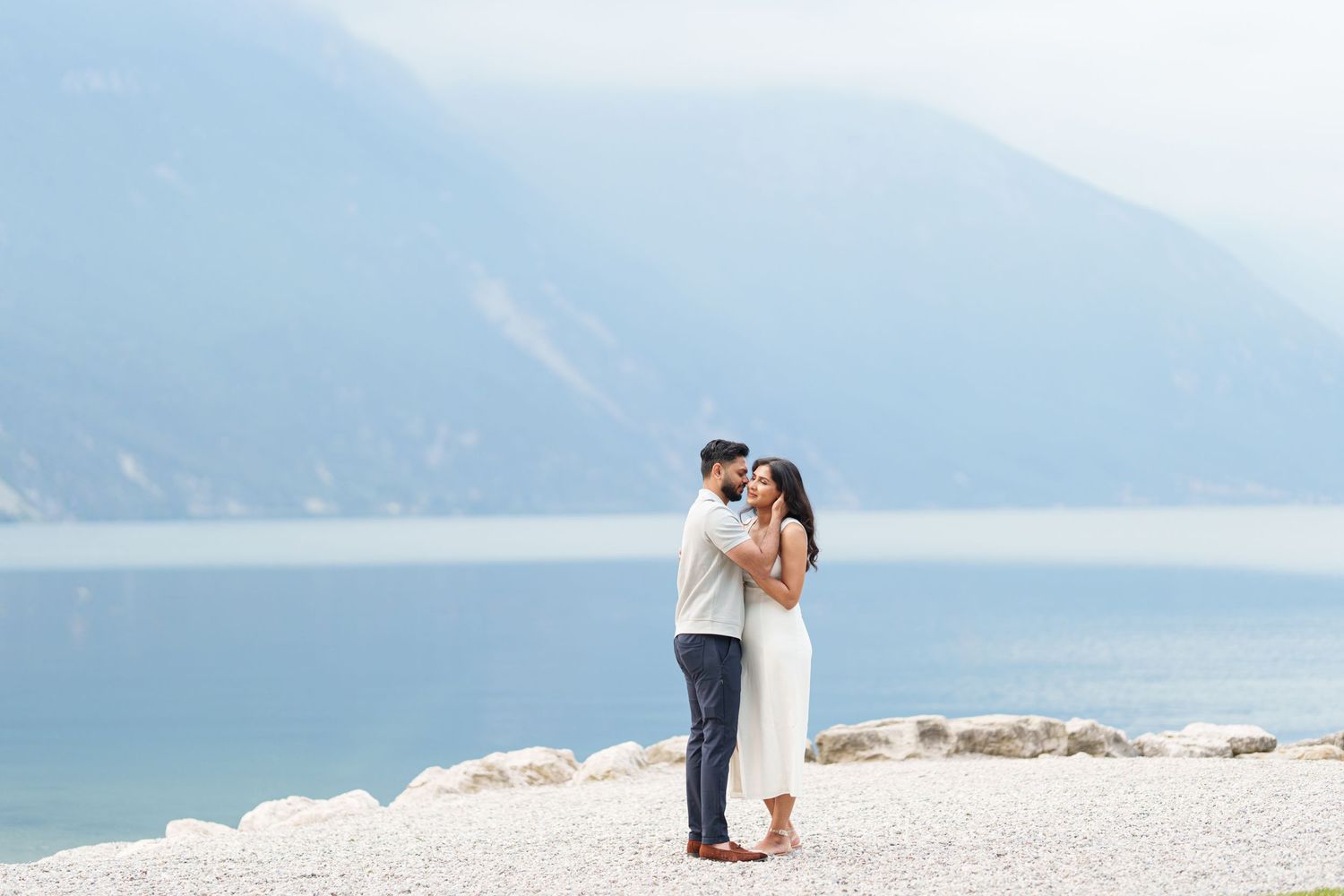 A couple embraces on a scenic mountain beach with blue water and misty peaks in the background.