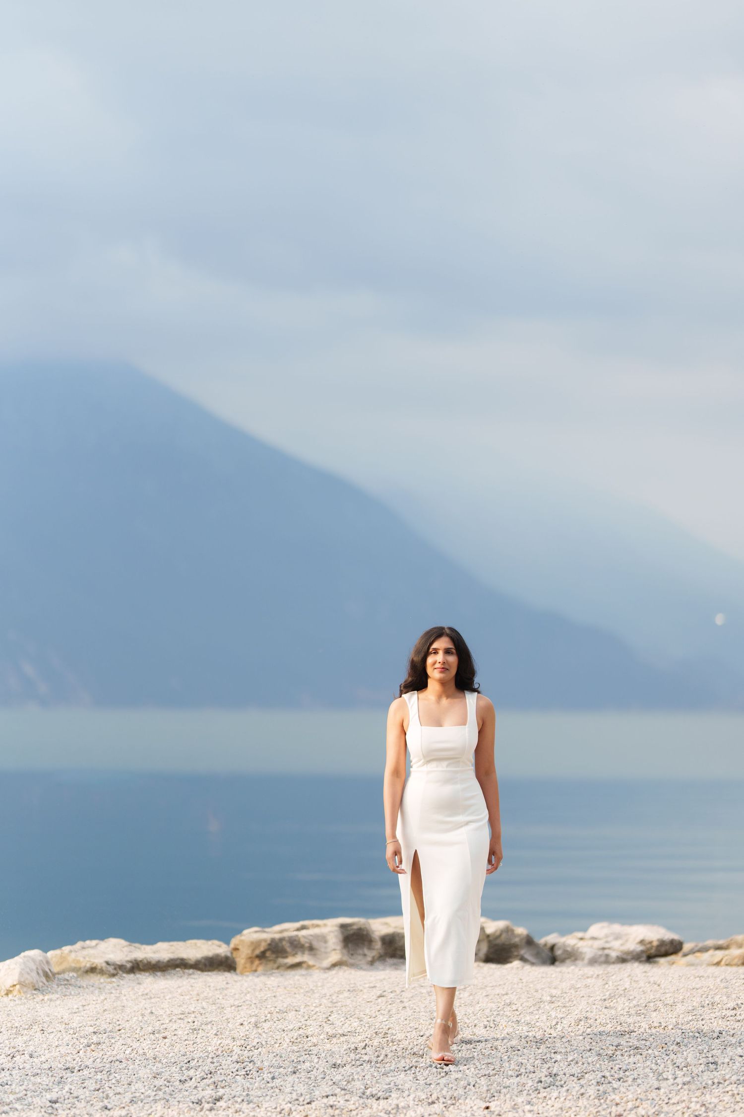 Person in white dress sits on rocky beach shore with misty mountains and lake in background creating serene scene.