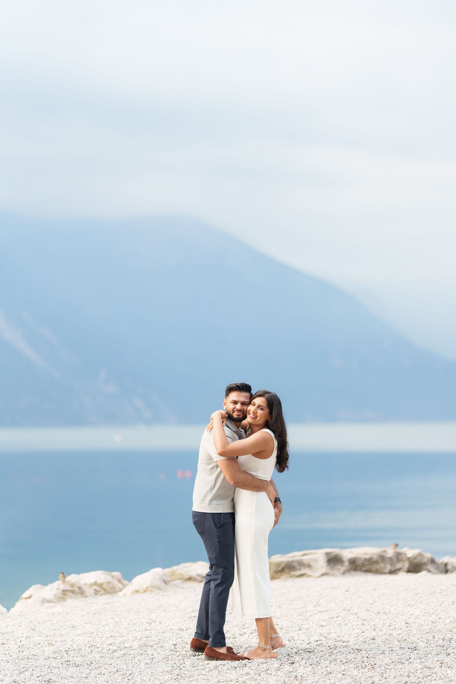 A couple embraces on a scenic beach with mountains and blue water in the background.