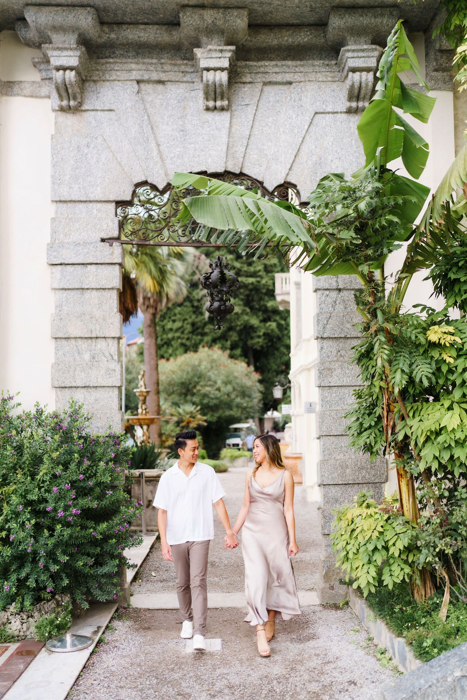 Two people walking through a stone archway surrounded by lush greenery and plants in a garden setting.