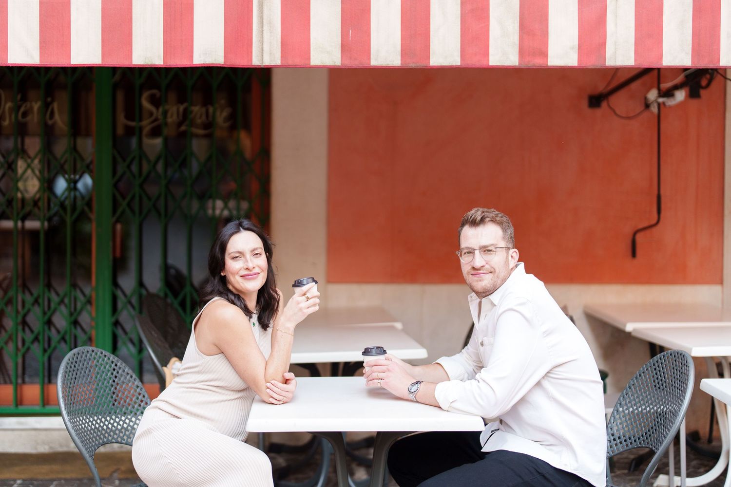 Two people in white shirts sit at an outdoor cafe table with red and white striped awning overhead.