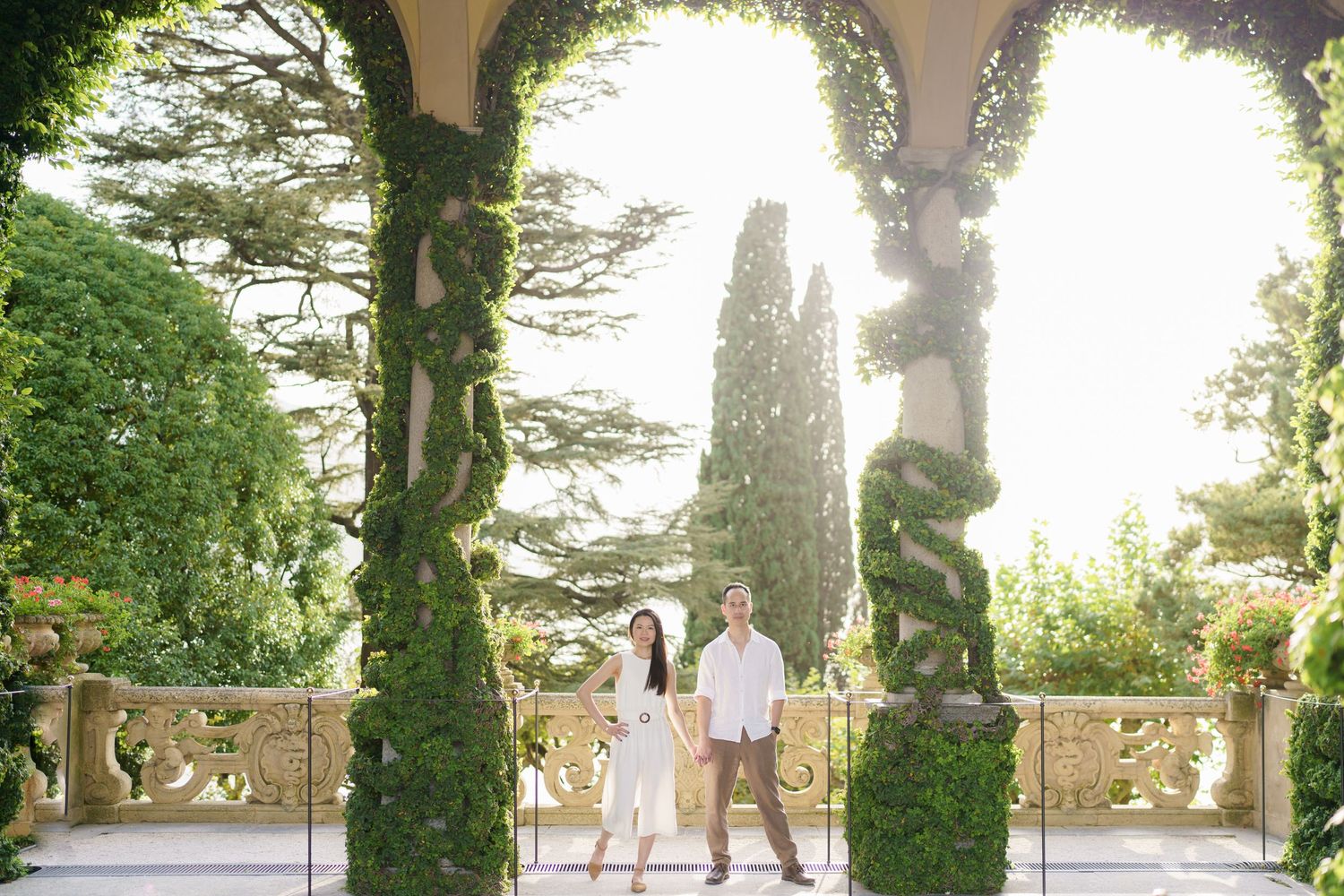 A couple stands between ivy-covered stone columns on an elegant Mediterranean terrace with cypress trees in the background.