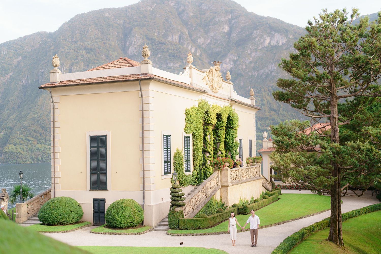 Elegant cream-colored villa with ornate details overlooking Lake Como in Italy, surrounded by manicured gardens and mountains.