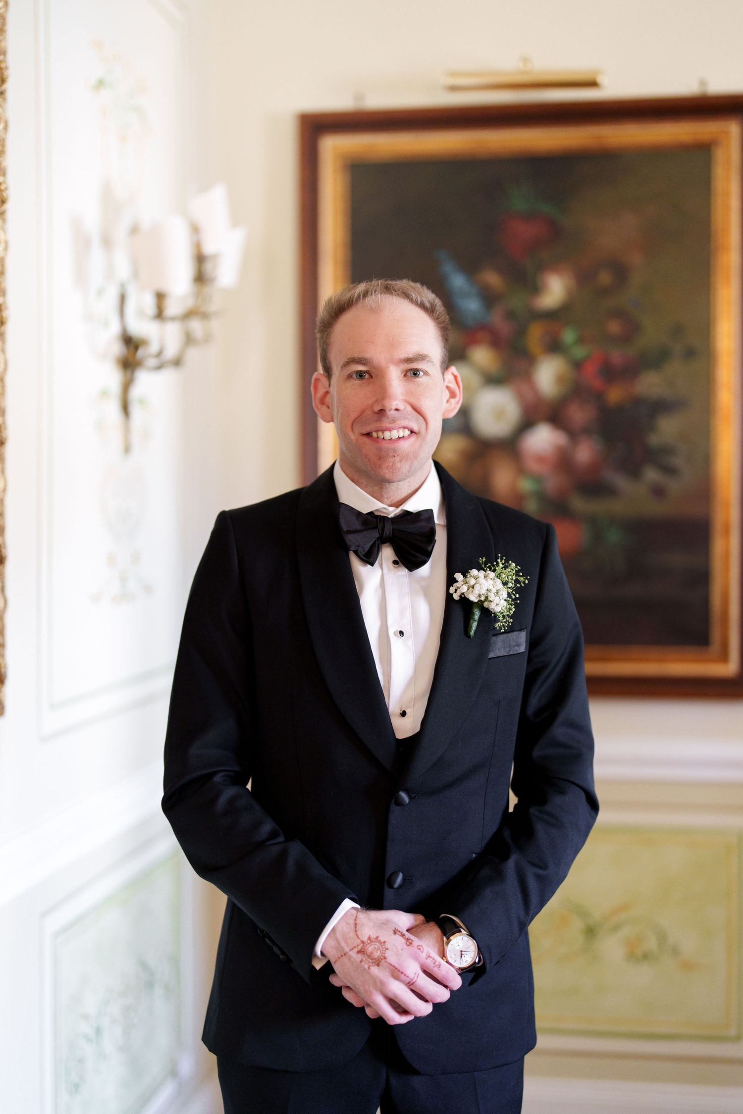 A person in formal attire smiles while standing in an elegant room with floral artwork.
