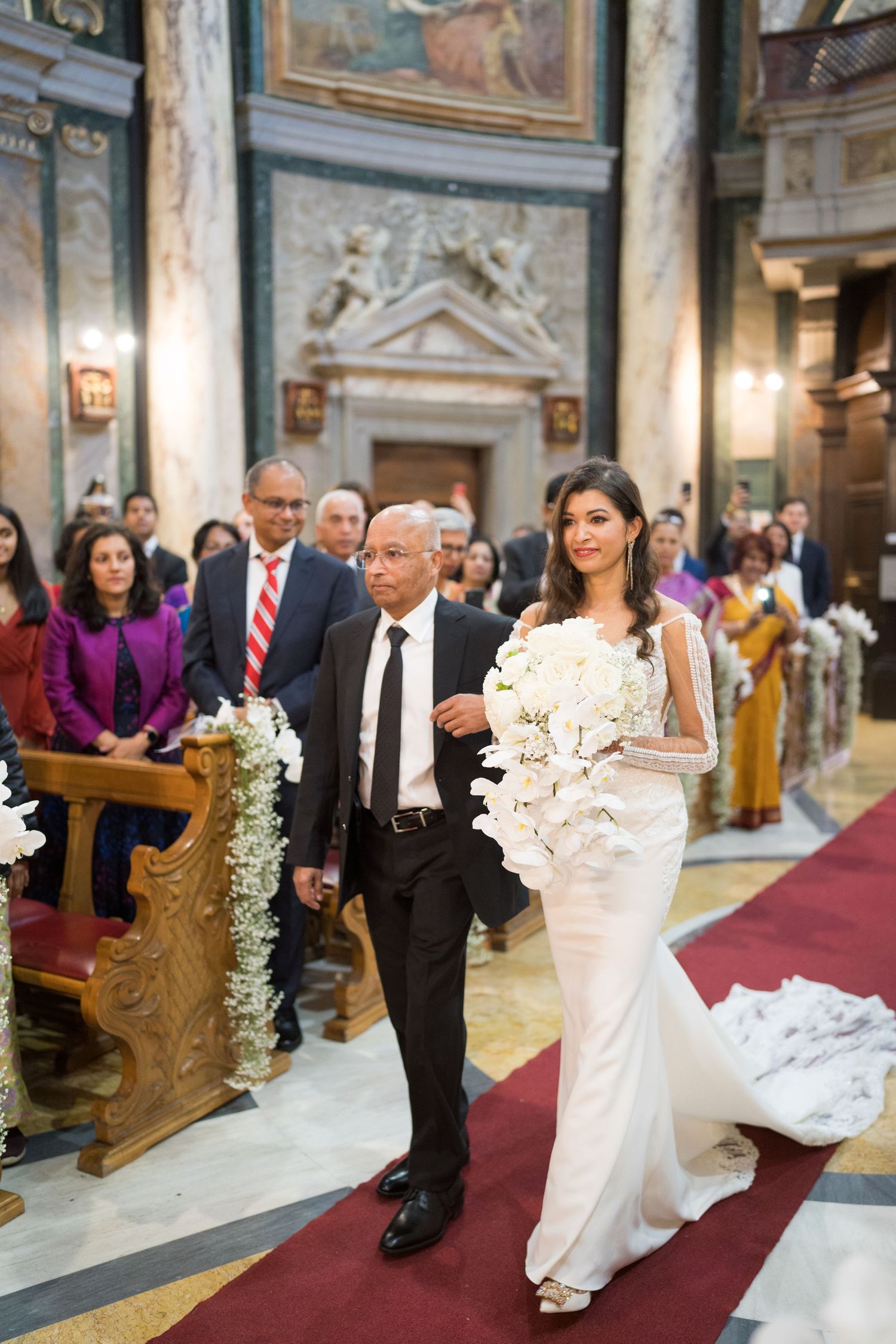 A wedding procession walking down a red-carpeted aisle in an elegant church with marble columns.