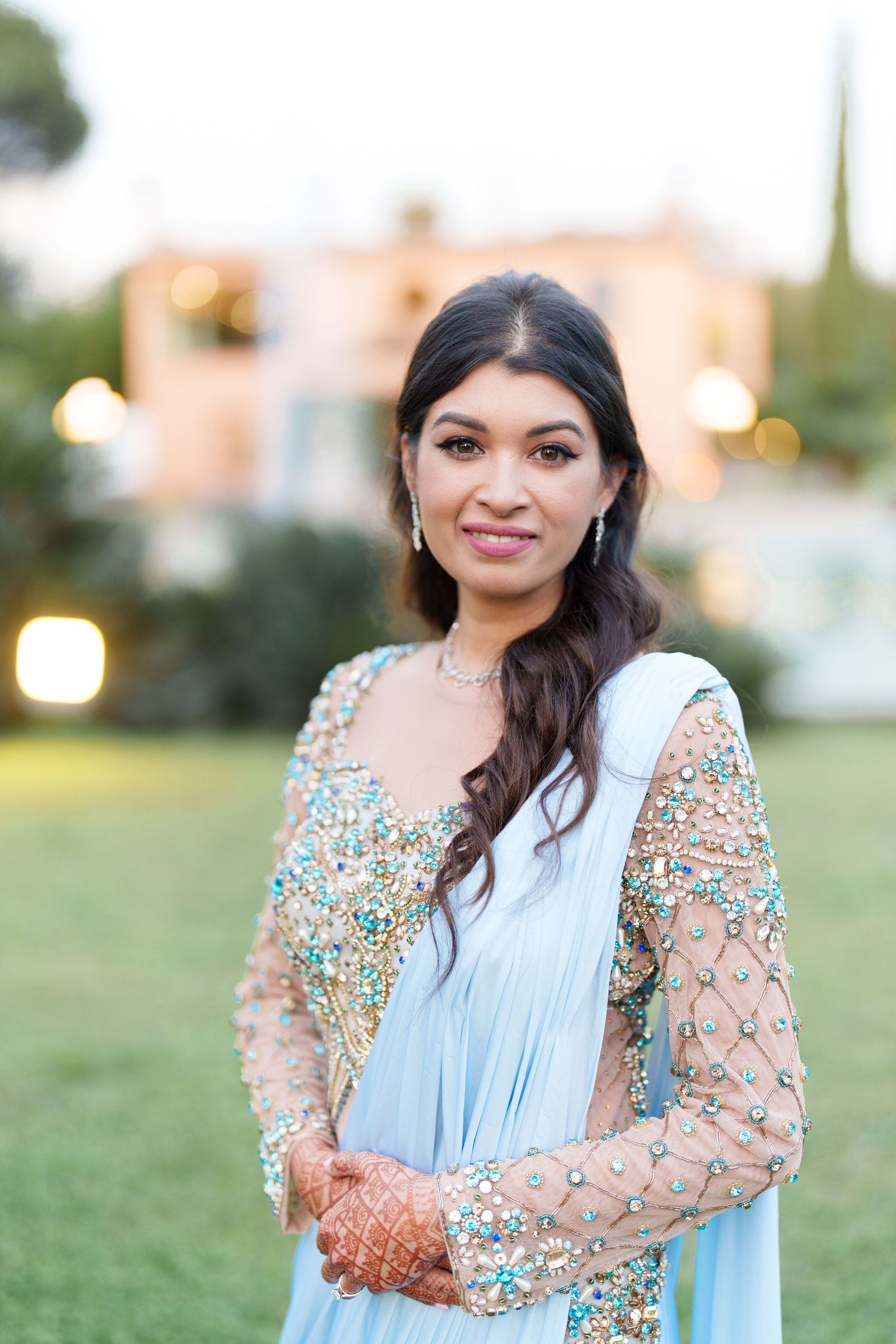 Woman in light blue dress with beaded details poses in garden setting at sunset.