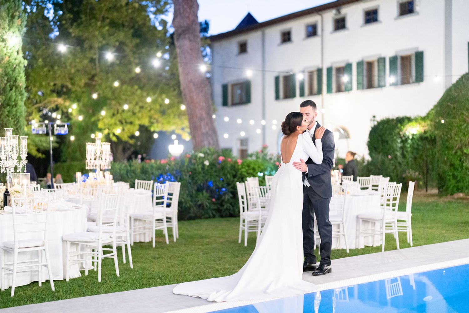 Evening wedding reception by illuminated pool with white chairs and string lights.