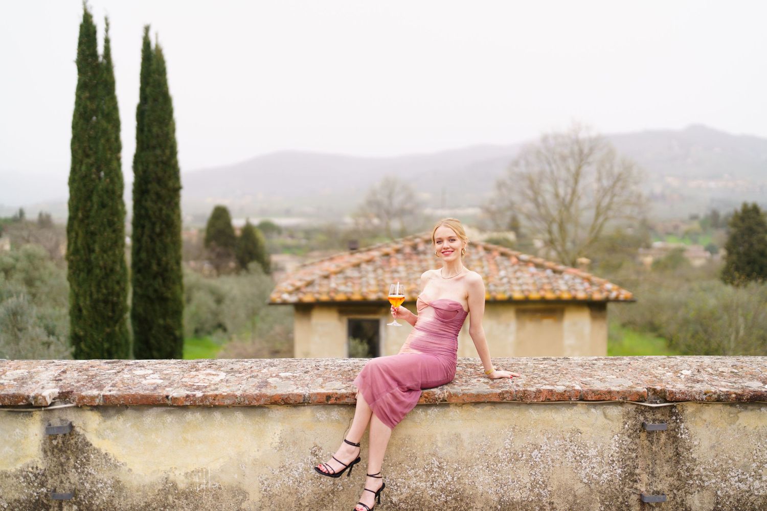 Stone wall and cypress trees frame a scenic Tuscan landscape view.