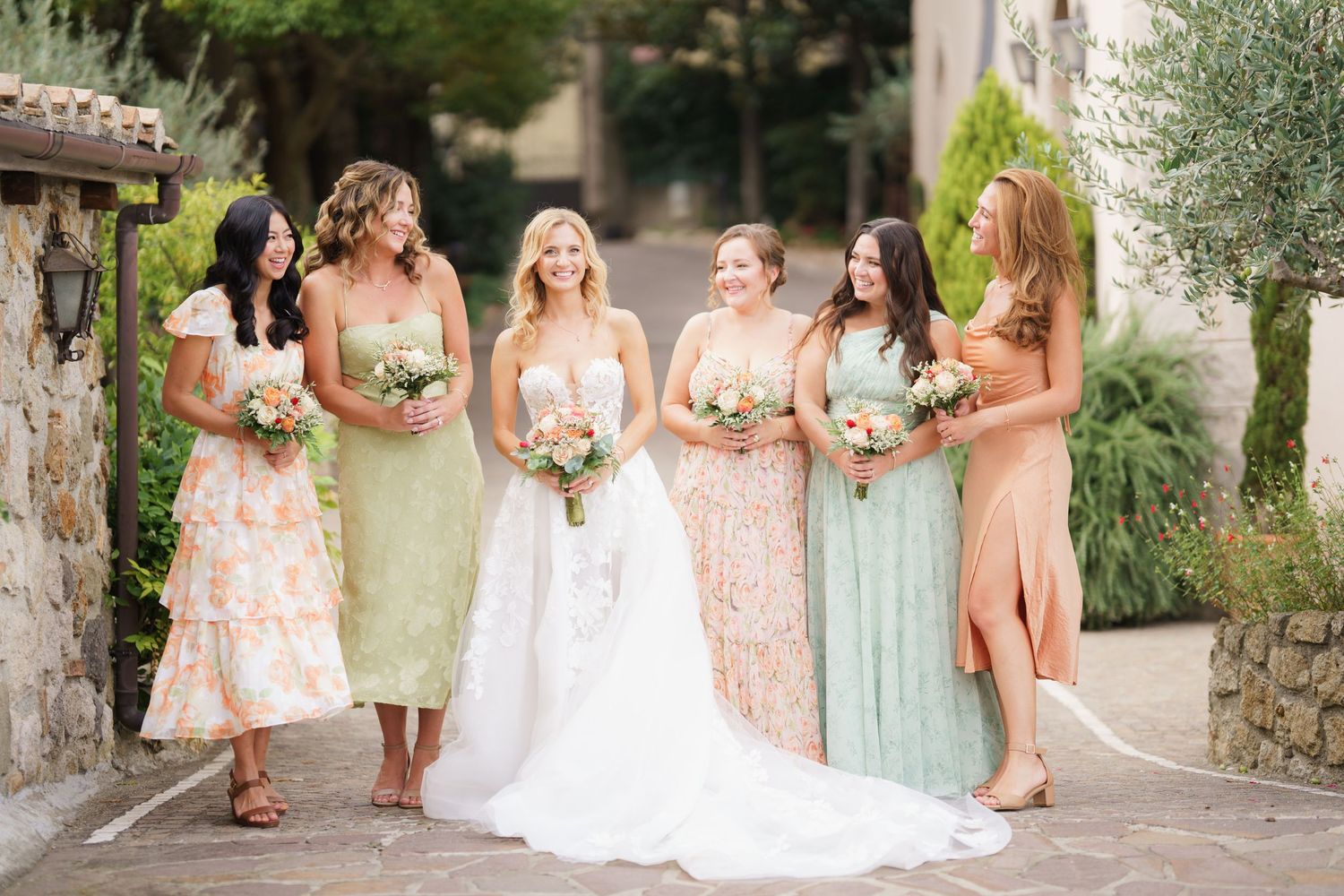 Wedding party in pastel dresses poses on stone steps surrounded by lush gardens.