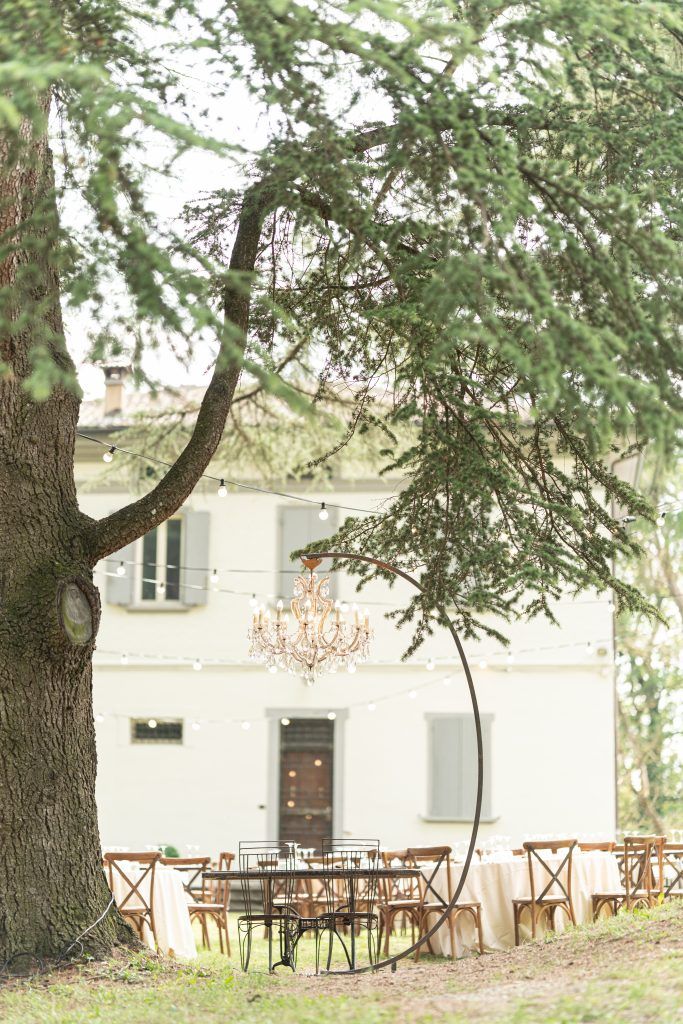 Wooden cross-back chairs arranged under large trees near a white building for an outdoor event.