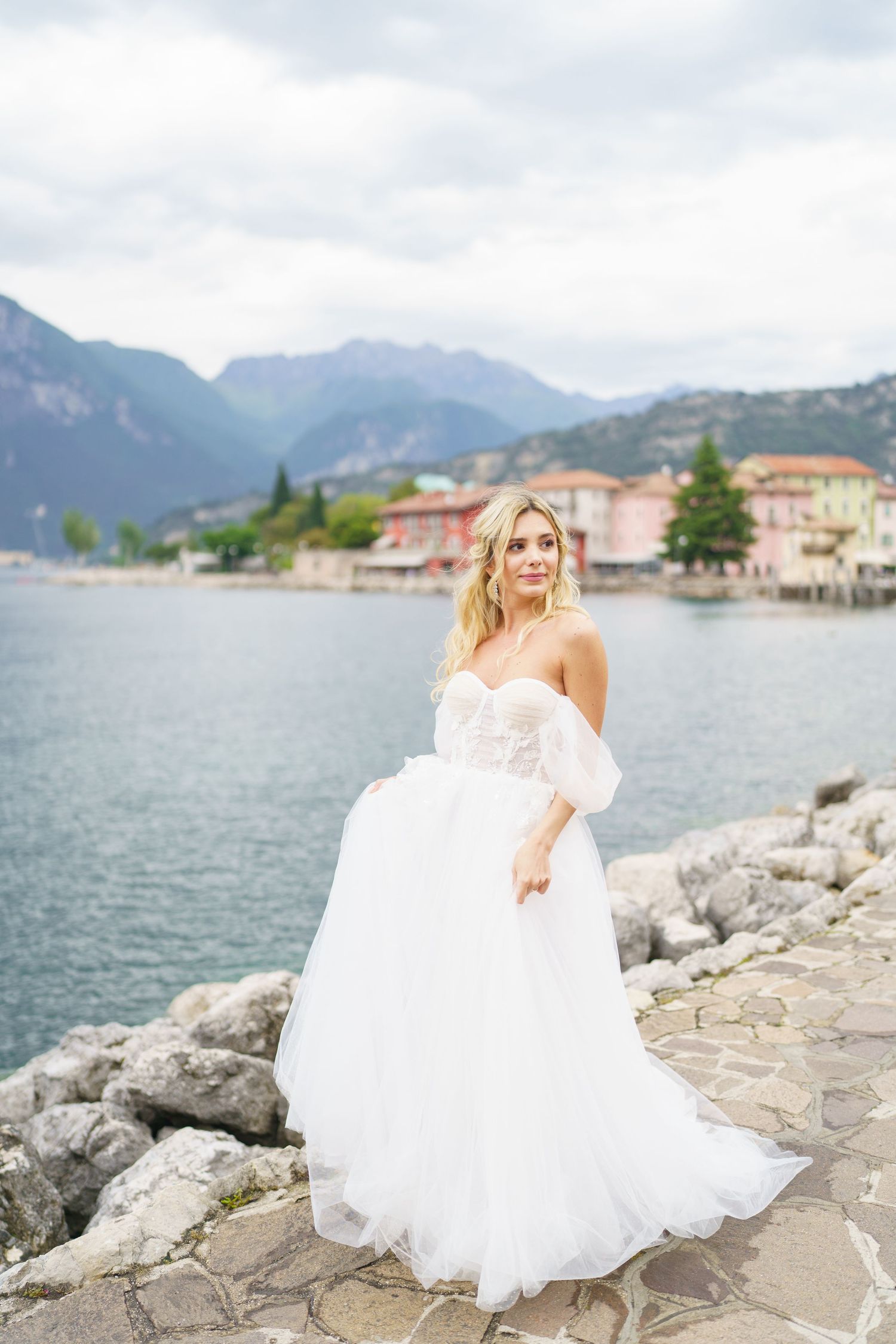 A bride in a flowing white wedding gown stands on rocky shores of Lake Como with Italian mountains and villas in background.