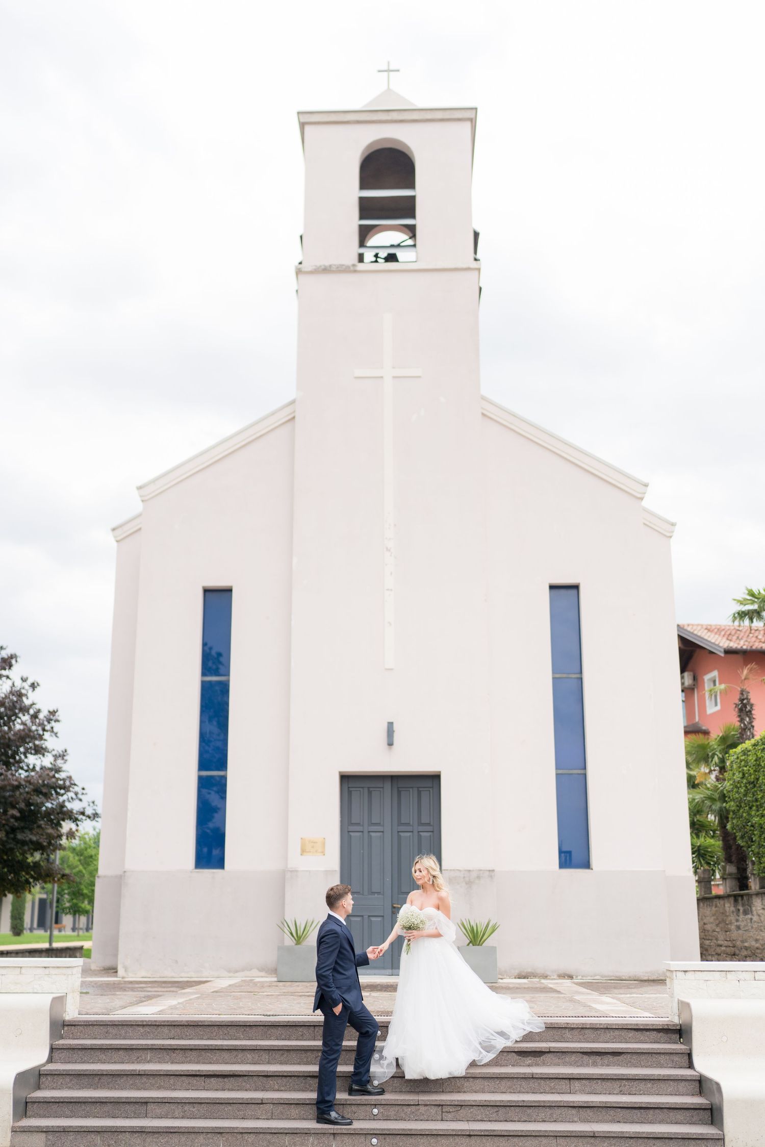 Wedding couple poses on steps in front of a white church with blue windows and gray doors.