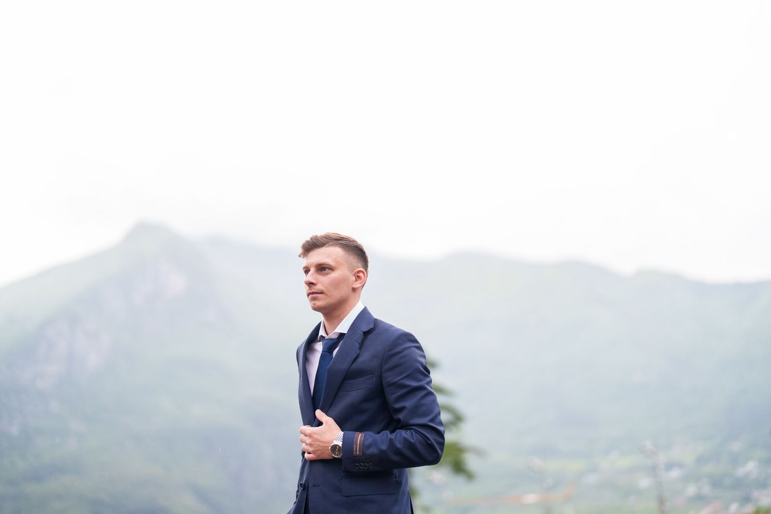 Professional in a navy suit stands on a mountain overlook with scenic mountain ranges in the misty background.
