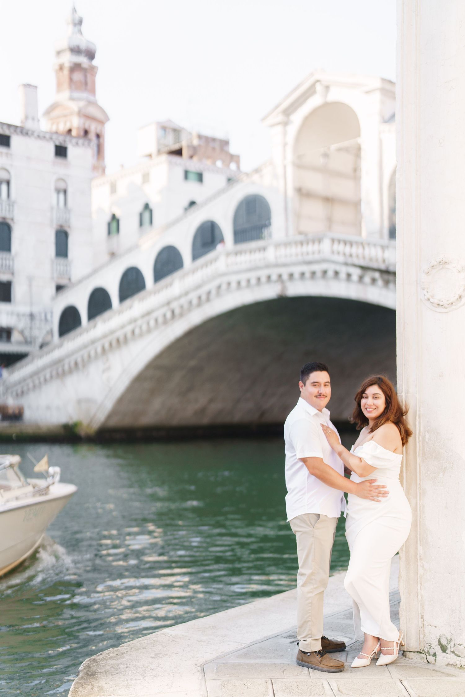 A couple in white outfits poses together near a historic stone bridge over a canal in Venice, Italy.