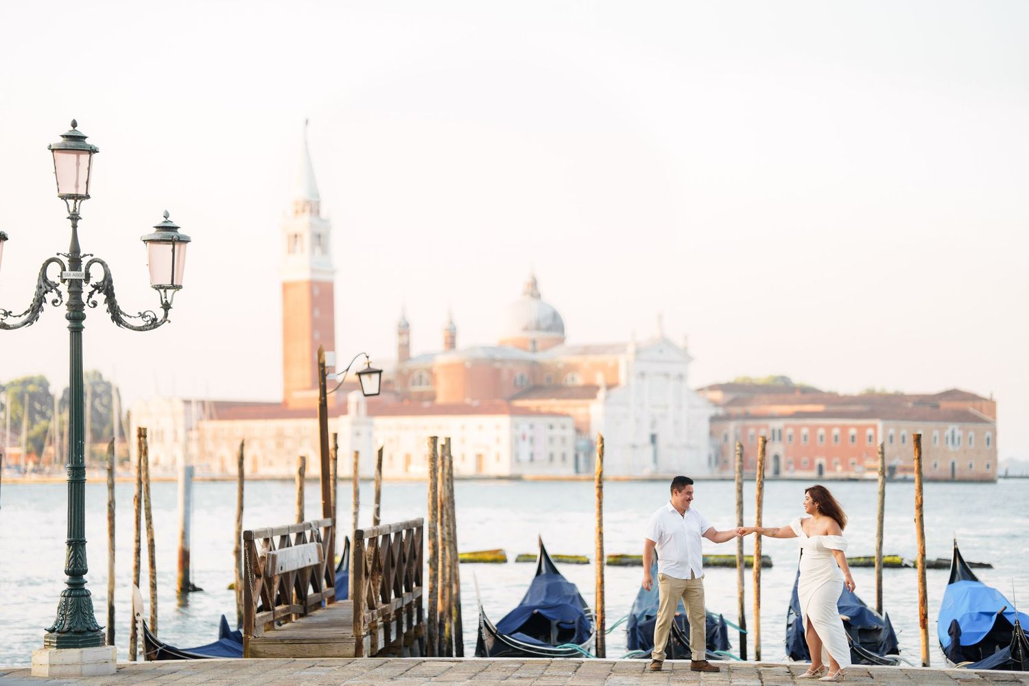 Gondolas moored along Venice waterfront with San Giorgio Maggiore church and ornate lamp post at sunset.