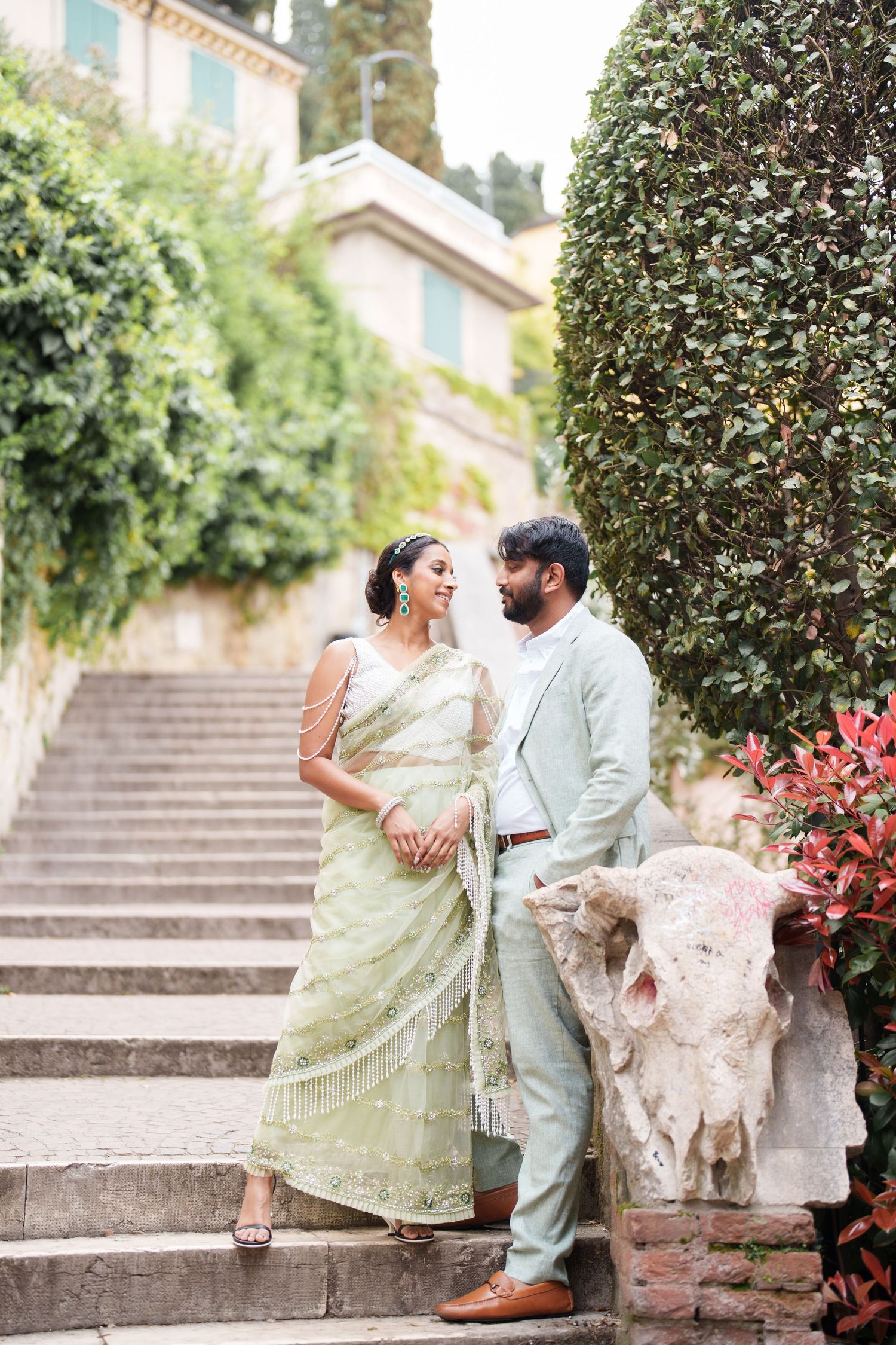 A couple in traditional Indian attire shares romantic moments on stone steps surrounded by greenery.