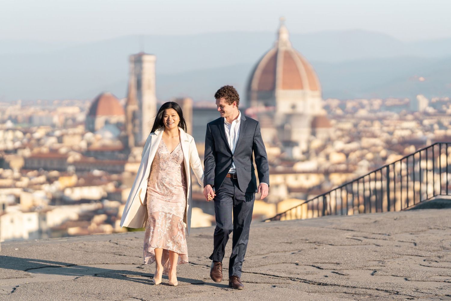 A couple walks along a scenic overlook in Florence, Italy with the iconic Duomo cathedral visible in the distance.