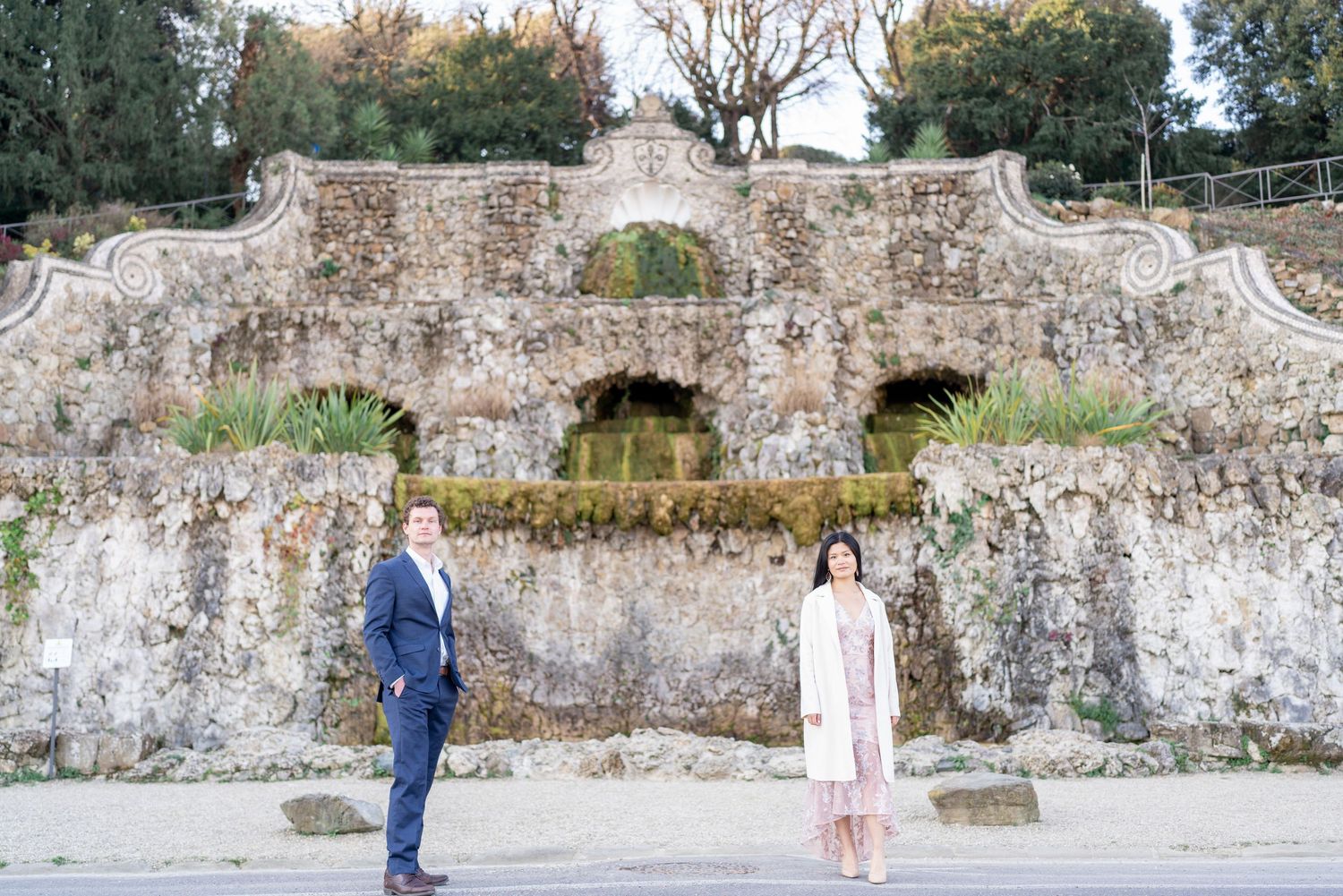 Two people stand in front of an ancient stone grotto wall with arches and weathered details in a garden setting.