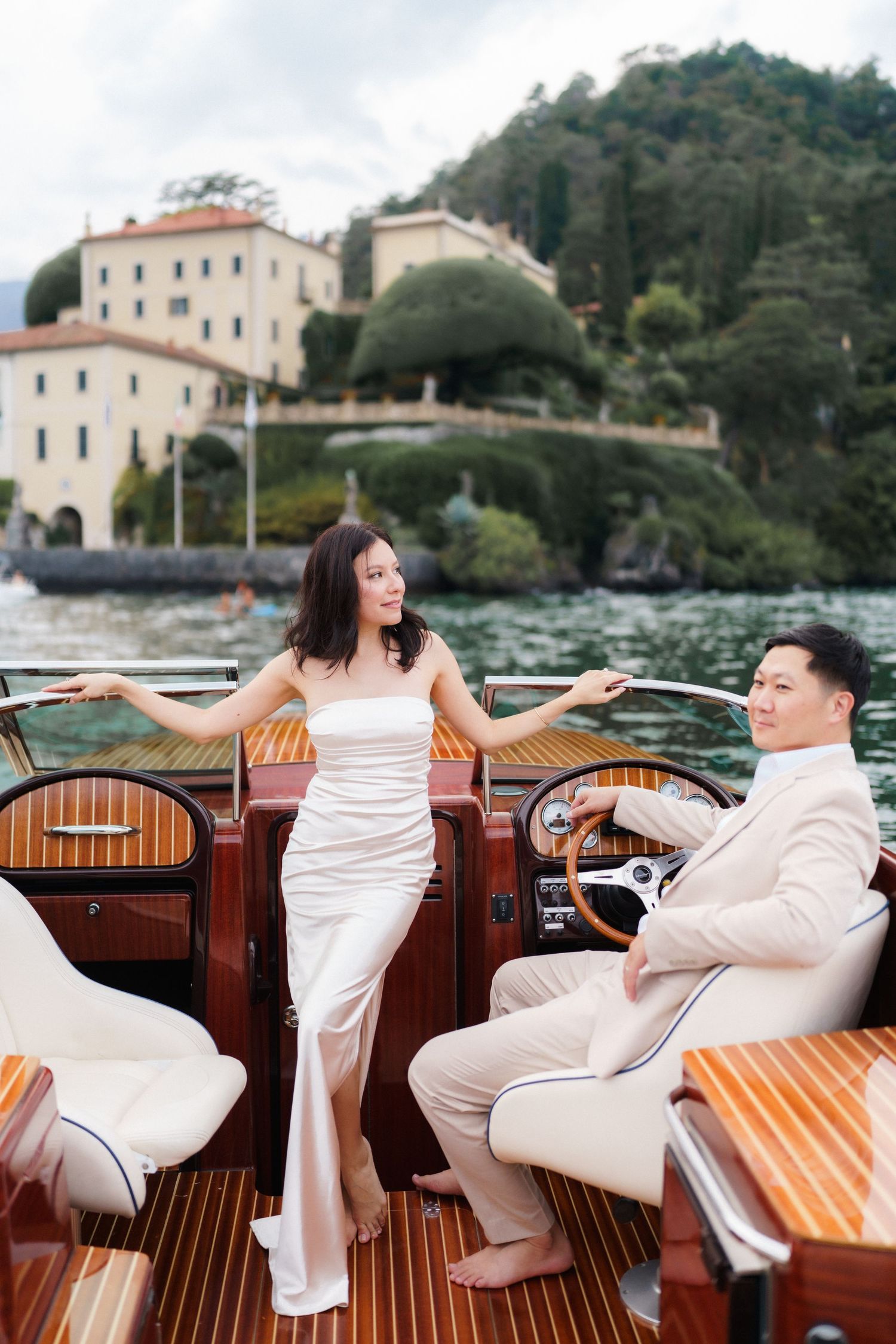 A romantic couple in white attire poses together on a wooden boat deck near coastal buildings and water.