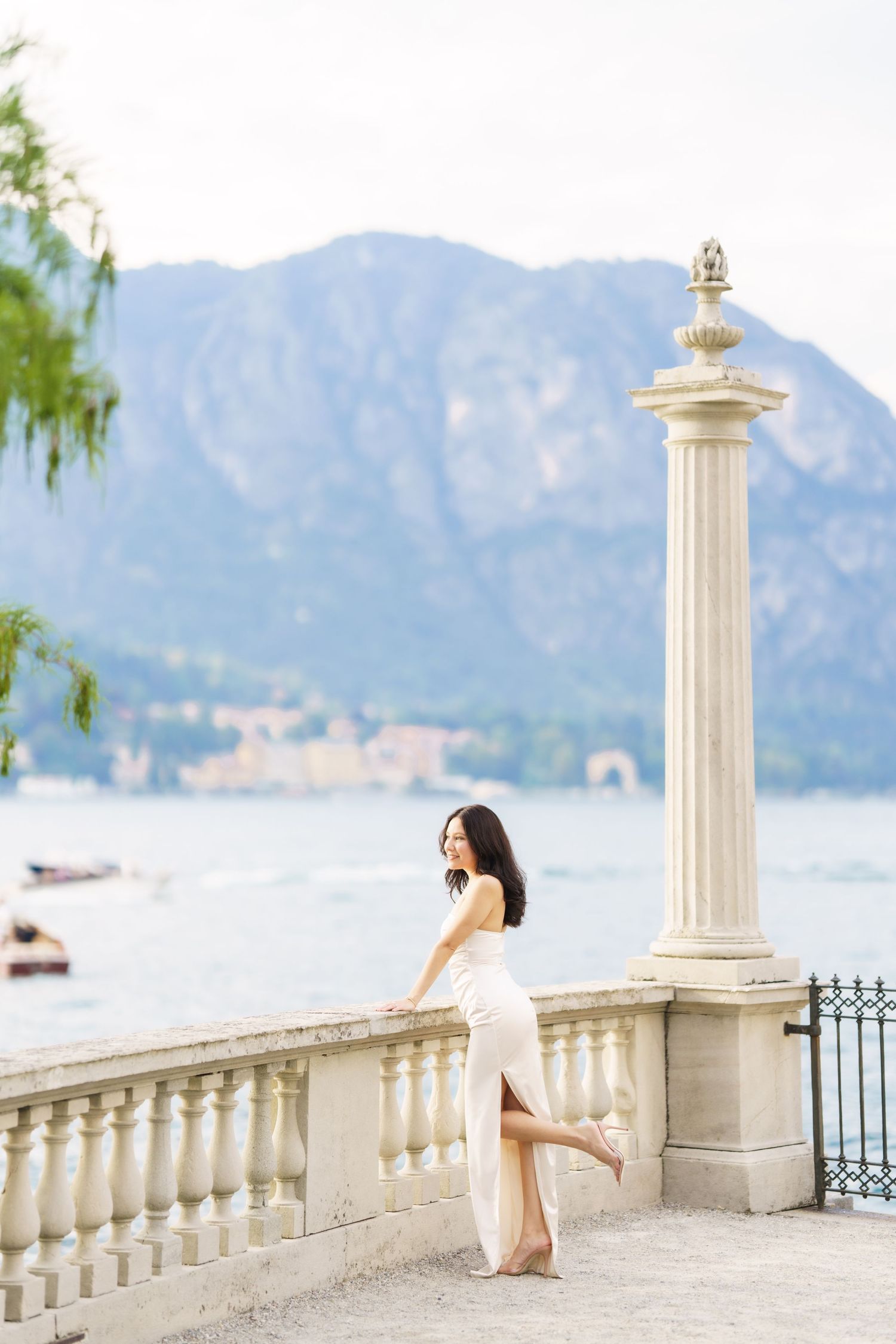A person in white stands by an ornate stone balustrade overlooking a scenic mountain lake view.