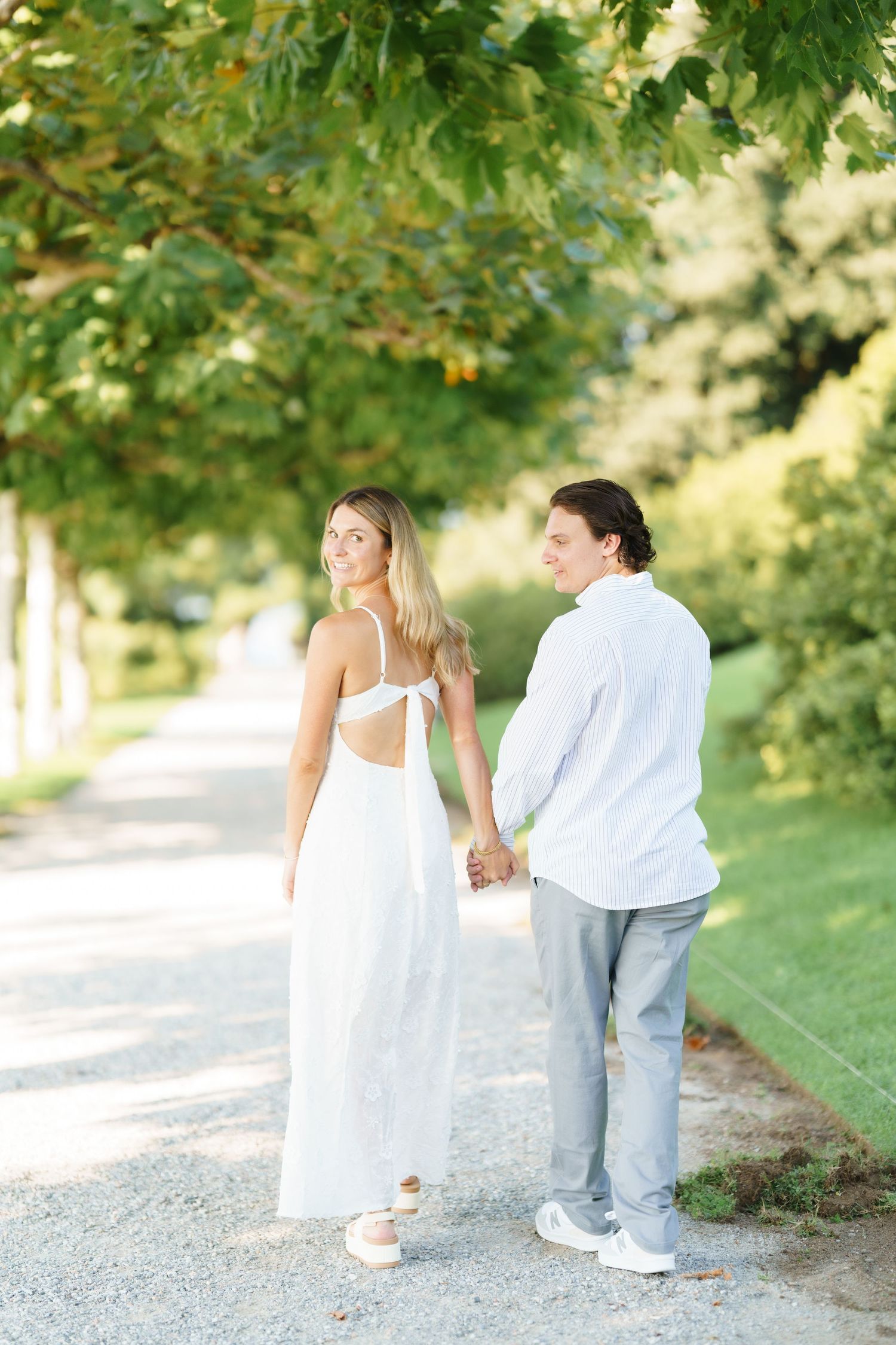 A couple in white attire walking together on a path surrounded by lush green trees on a sunny day.