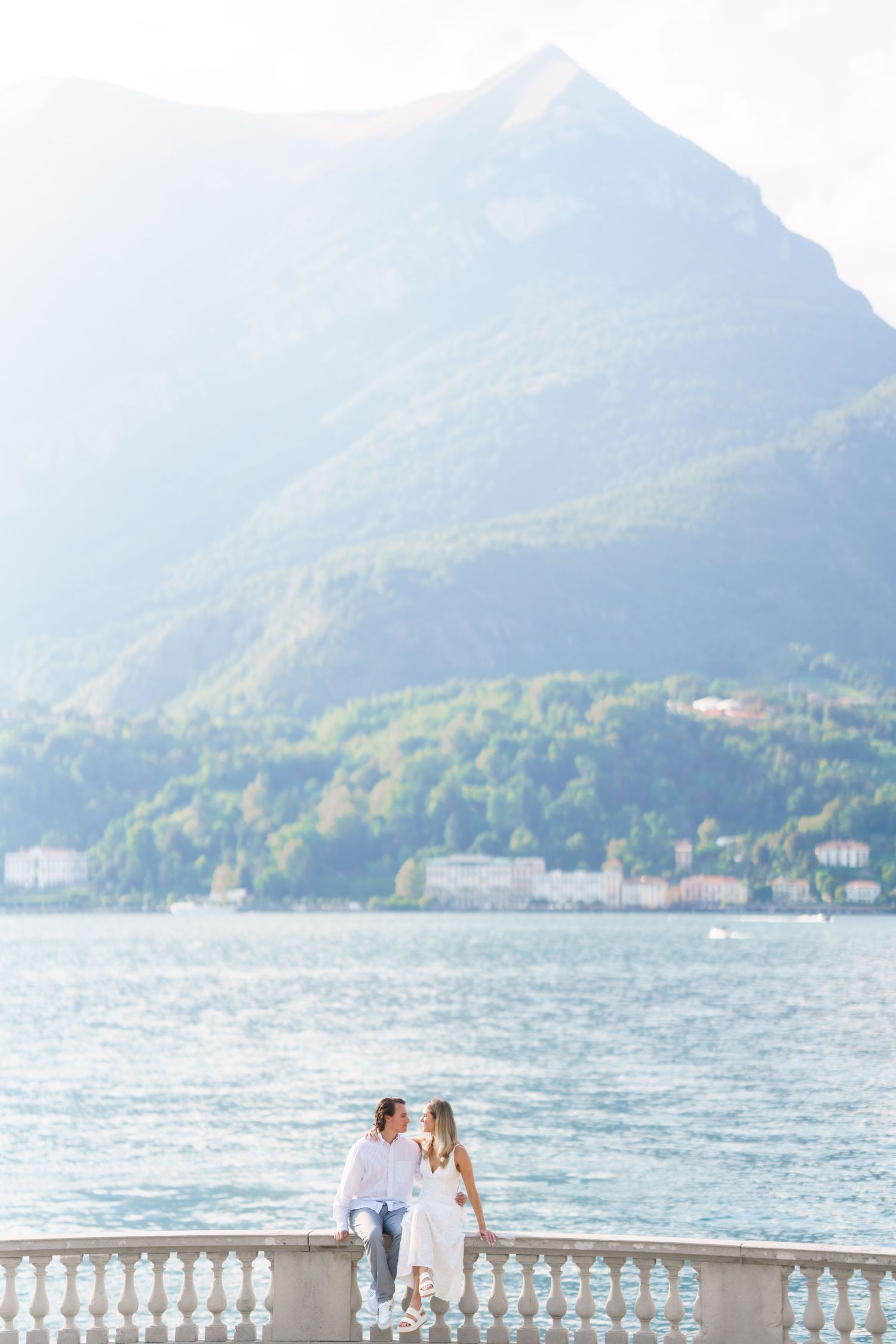 A couple shares a romantic moment on a lakeside terrace with mountains and water in the background.