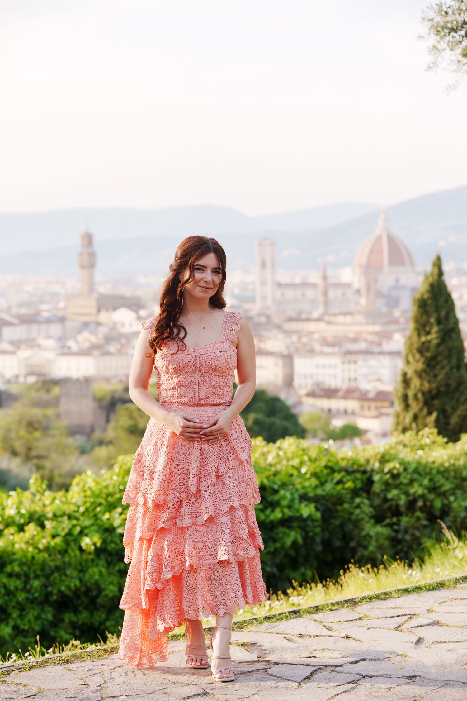 A woman in a pink lace dress poses at Piazzale Michelangelo overlooking the Florence skyline with the Duomo visible.