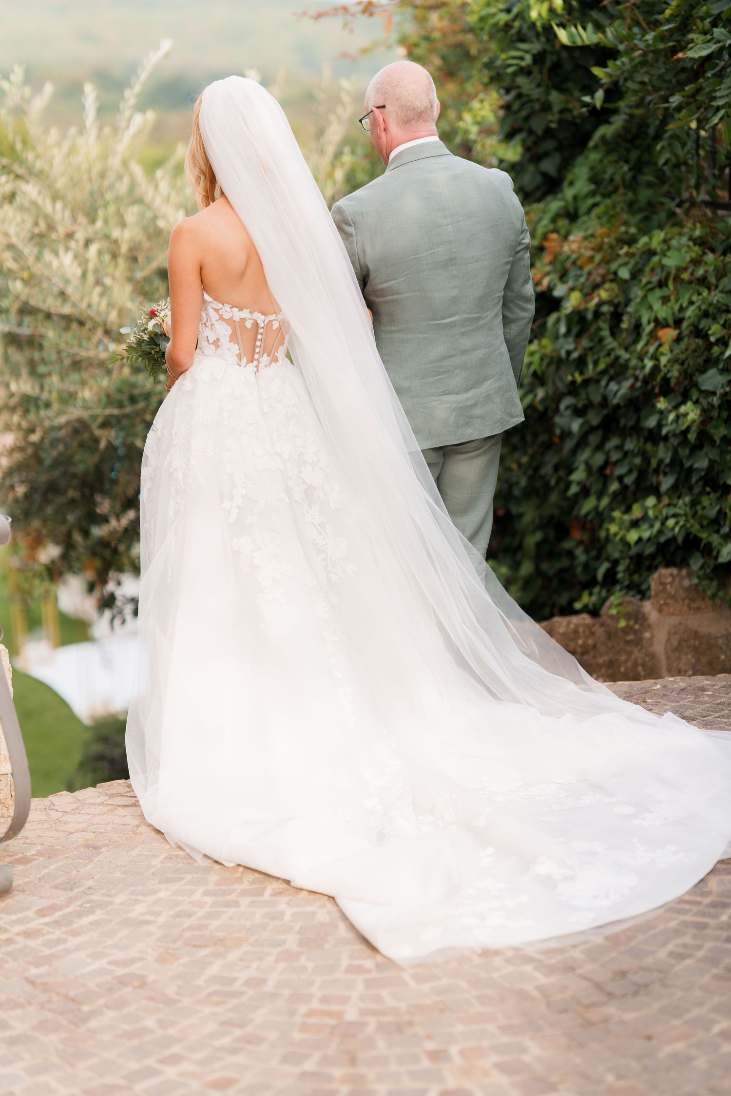 Wedding couple walking together on stone path with flowing white dress trailing behind them.