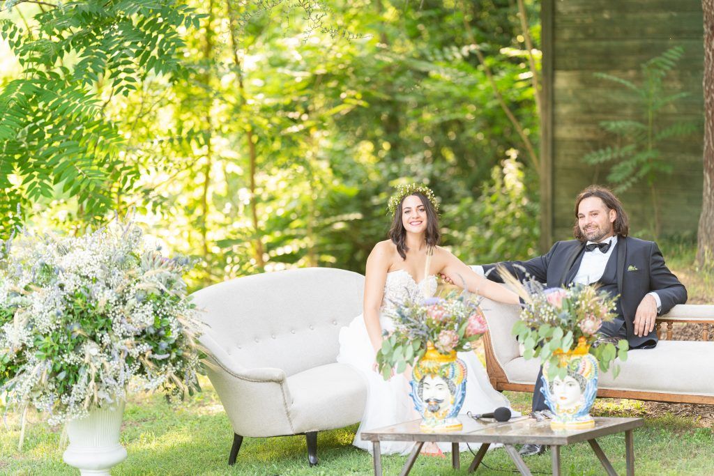 Garden seating area with white vintage furniture arranged on a lawn surrounded by lush greenery.