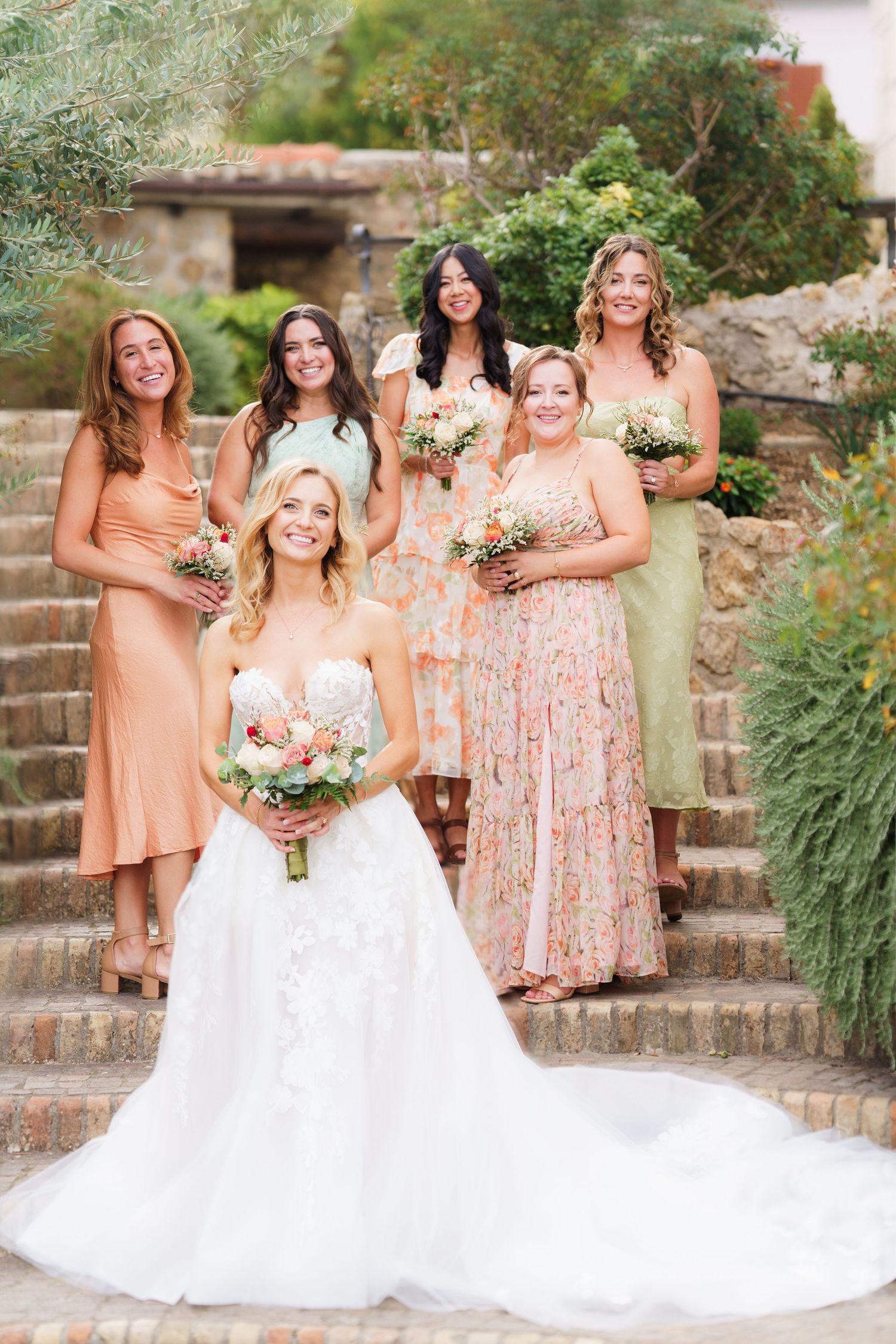 Bridal party wearing soft peach and mint dresses gather on rustic stone staircase.