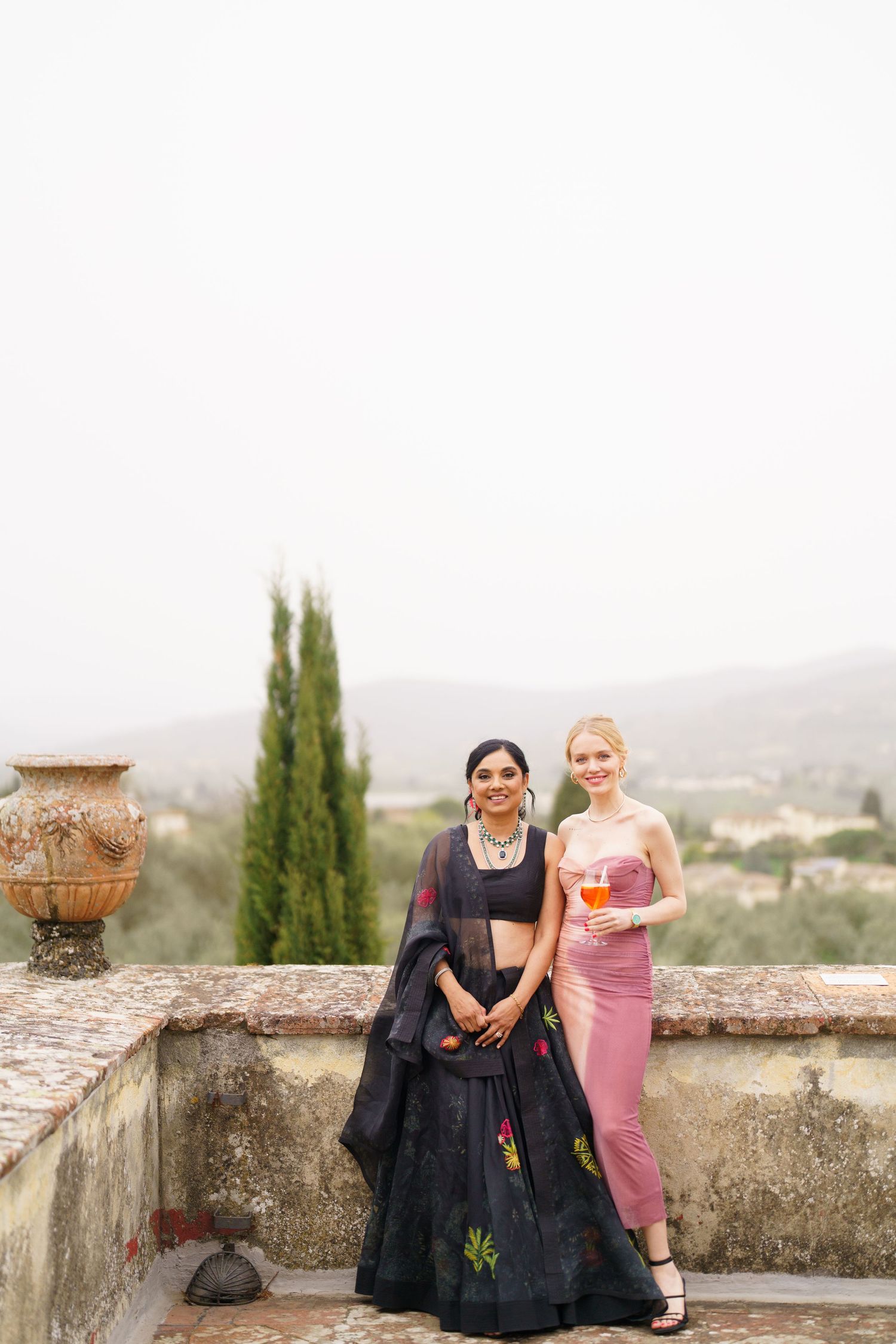 Two people in contrasting outfits sitting together on ancient stone wall with mountain vista.