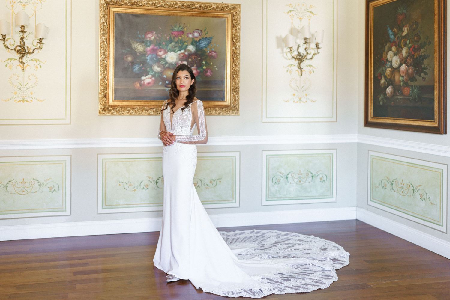 Bride in flowing white gown poses in luxurious room with ornate gold frames and crystal chandeliers.