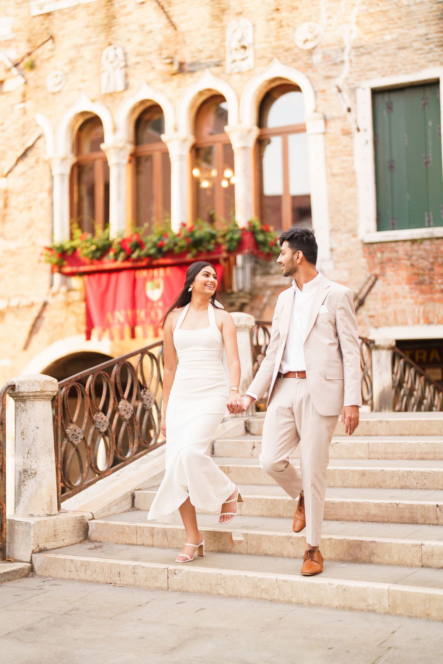 Couple in white outfits walking along stone steps of a historic European building with arched windows and red decor.