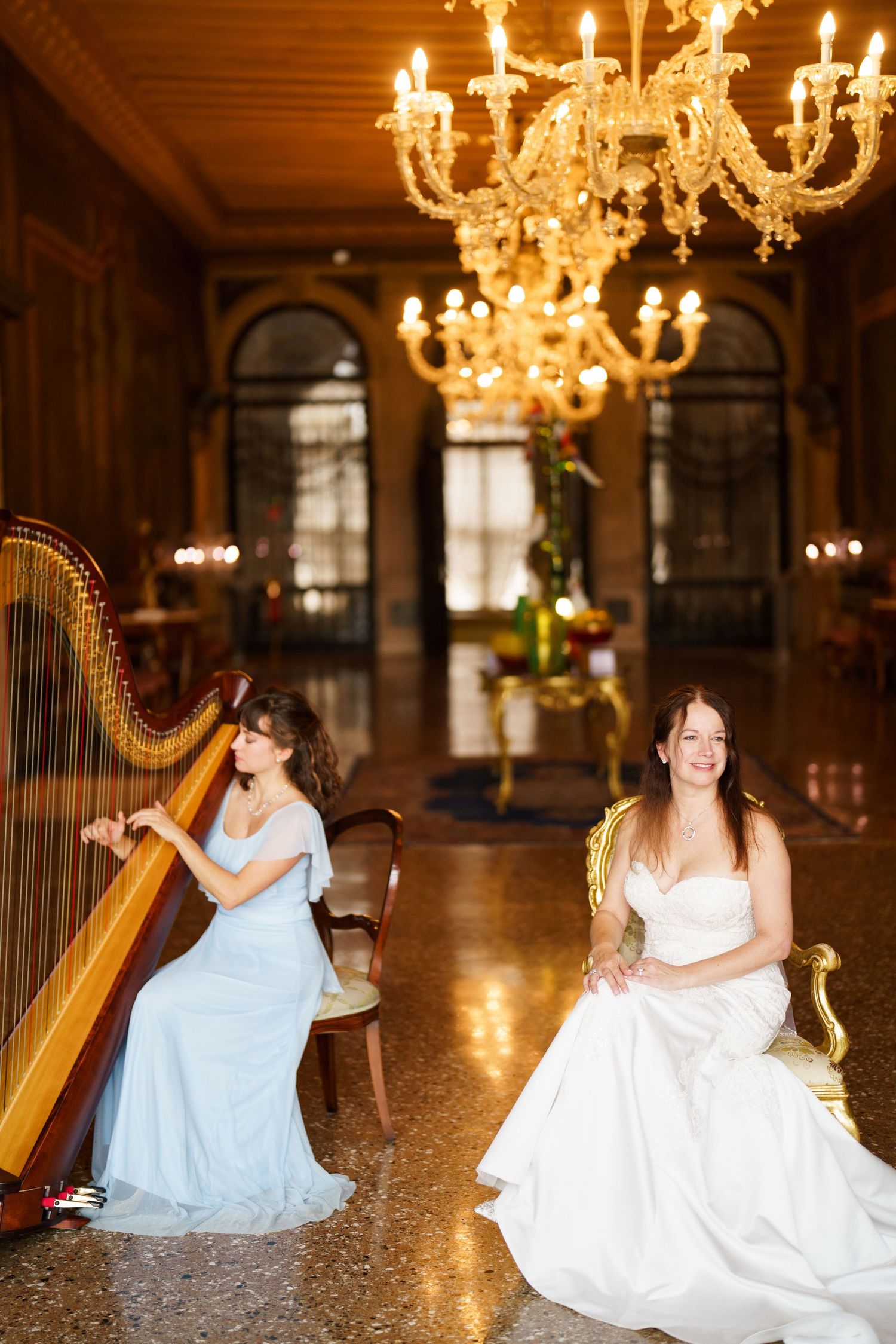 Two musicians in formal dresses play elegant harps in a grand ballroom with crystal chandeliers overhead.