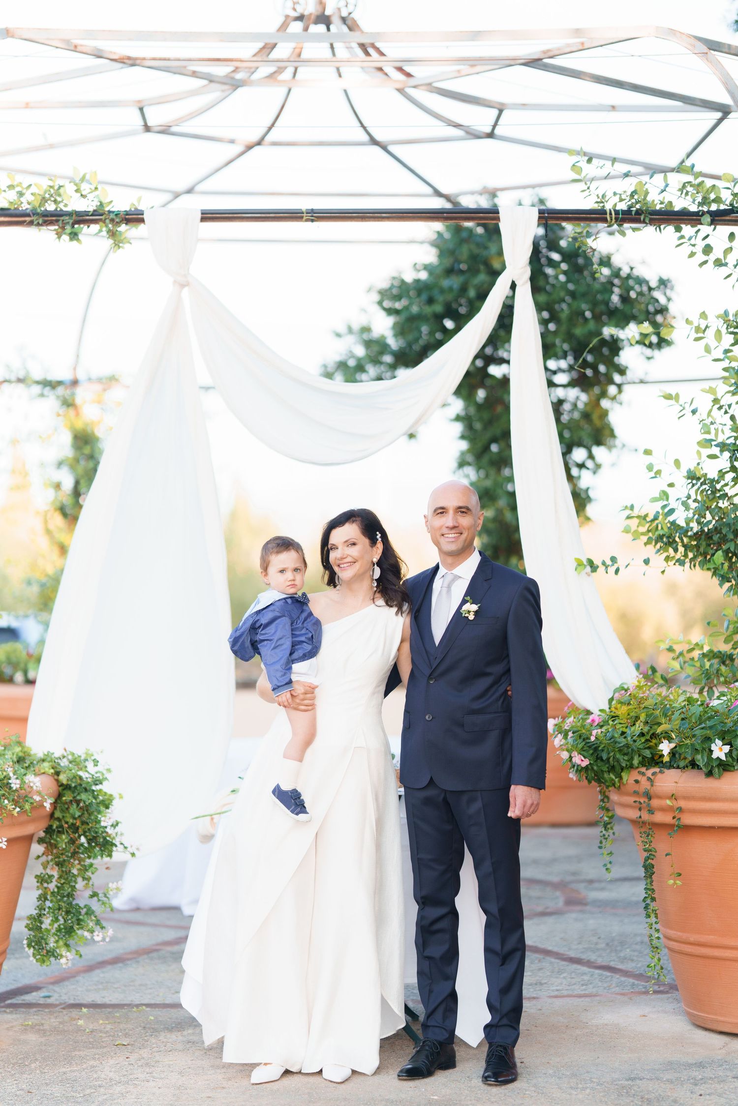 Garden wedding scene with white fabric draping and decorative potted plants at sunset.