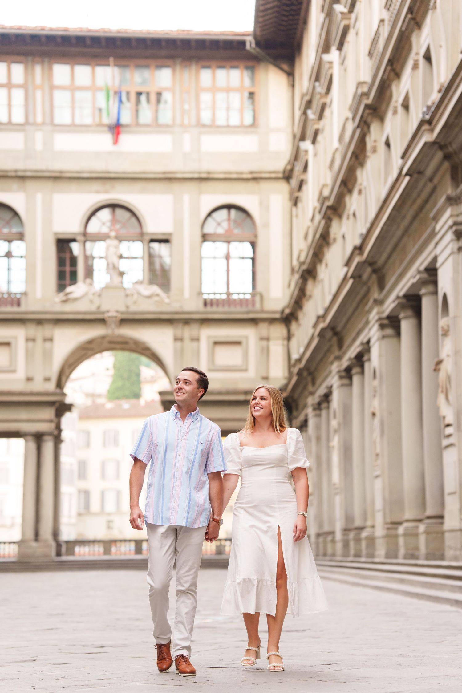 A couple in light summer attire walks hand in hand through a grand European architectural corridor.