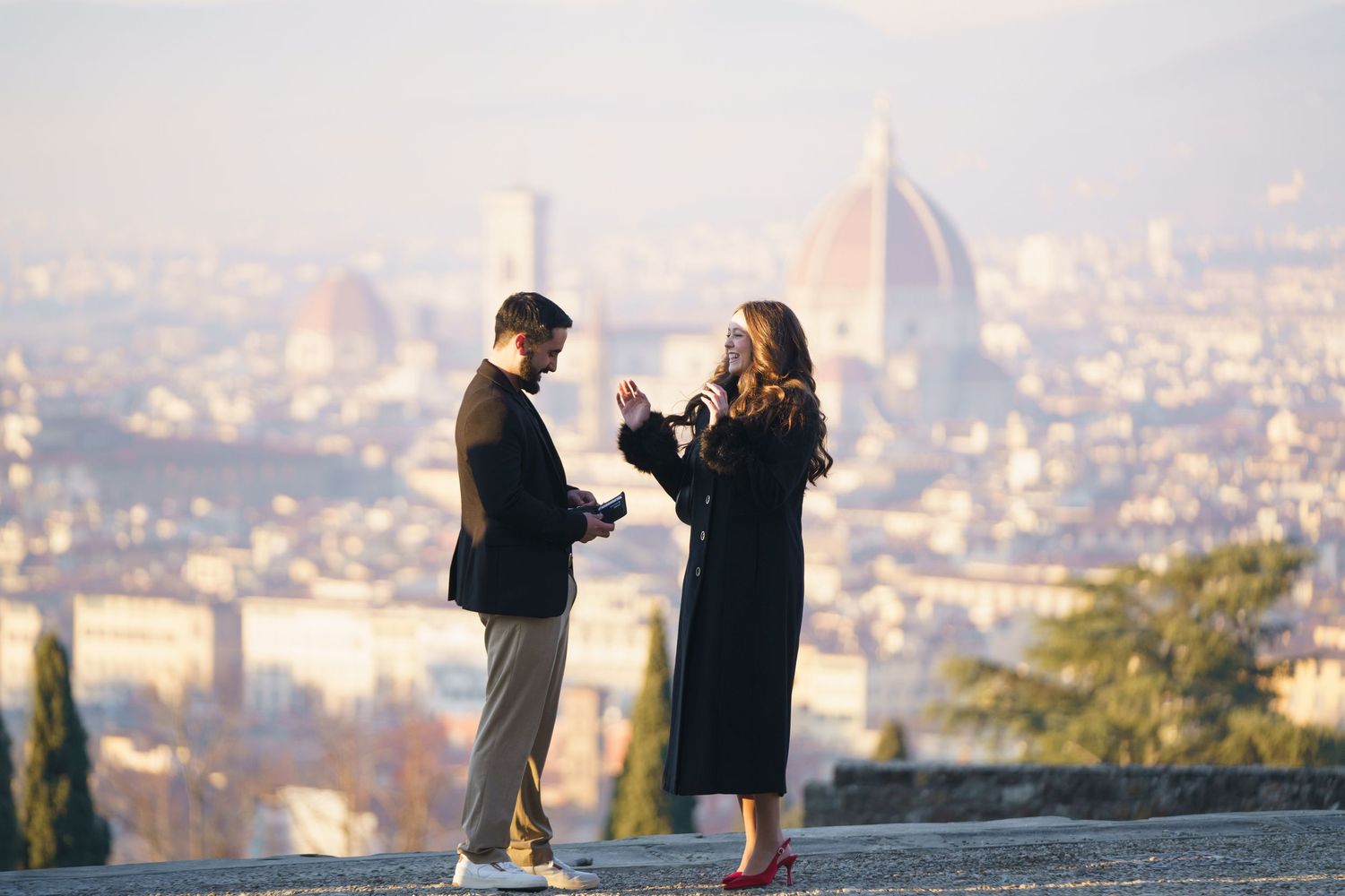 A couple stands on a scenic hilltop overlooking a city during a romantic sunset proposal moment.