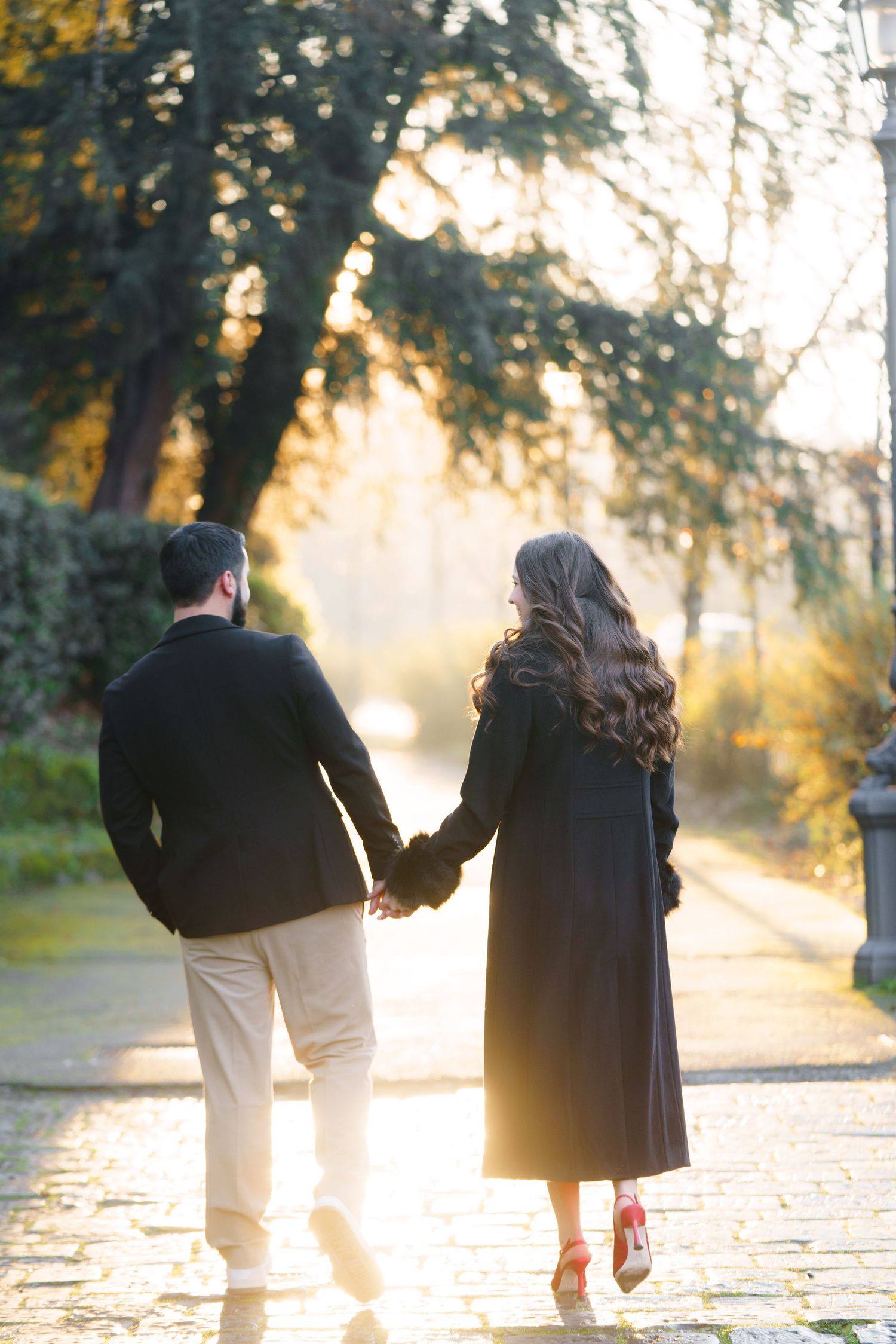 A couple walks hand in hand along a sunlit path during golden hour as sunbeams filter through autumn trees.